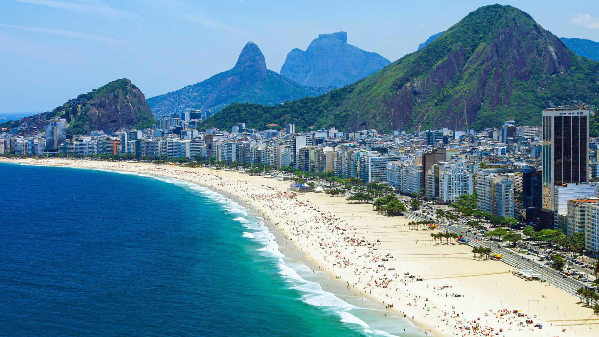  An aerial or elevated view of the curved Copacabana beach in Rio de Janeiro, Brazil, showing the wide white sand and the vast Atlantic Ocean. The famous black and white wave patterned promenade runs parallel to the beach, backed by a dense line of high rise apartment buildings and hotels, with mountains visible in the distance.