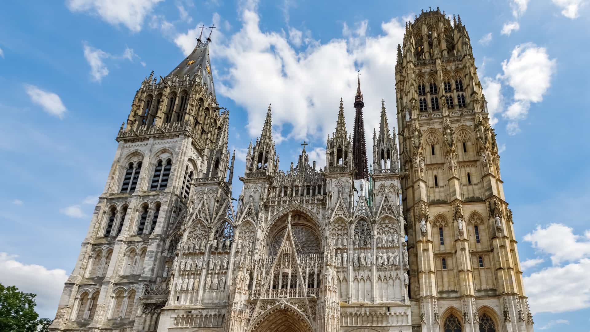  A magnificent, detailed view of the highly ornate, asymmetrical west facade of the Cathédrale Notre Dame de Rouen (Rouen Cathedral), a masterpiece of Gothic and Flamboyant architecture. The image focuses on the intricate stone carvings, numerous statues, and rose window above the main portal, with the flanking, mismatched Saint Romain Tower and the richly decorated Butter Tower (Tour de Beurre) prominent against the sky.