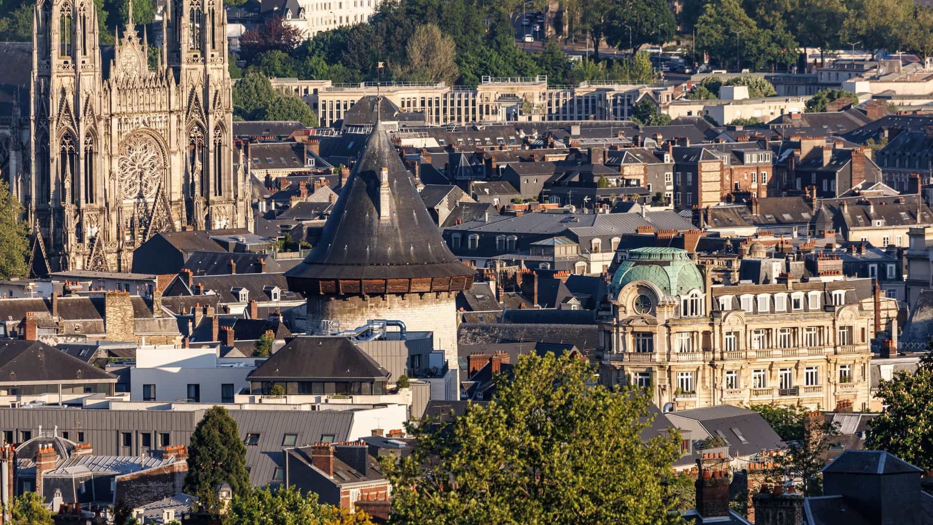  An aerial view of the city of Rouen, Normandy, France, showcasing a dense urban landscape of buildings and streets. The scene features the cylindrical stone keep of the historic Rouen Castle, officially known as the "Tour Jeanne d'Arc" (Joan of Arc Tower), prominent in the foreground, with the large Gothic structure of Saint Ouen Church and the Bonsecours Basilica visible on the horizon.