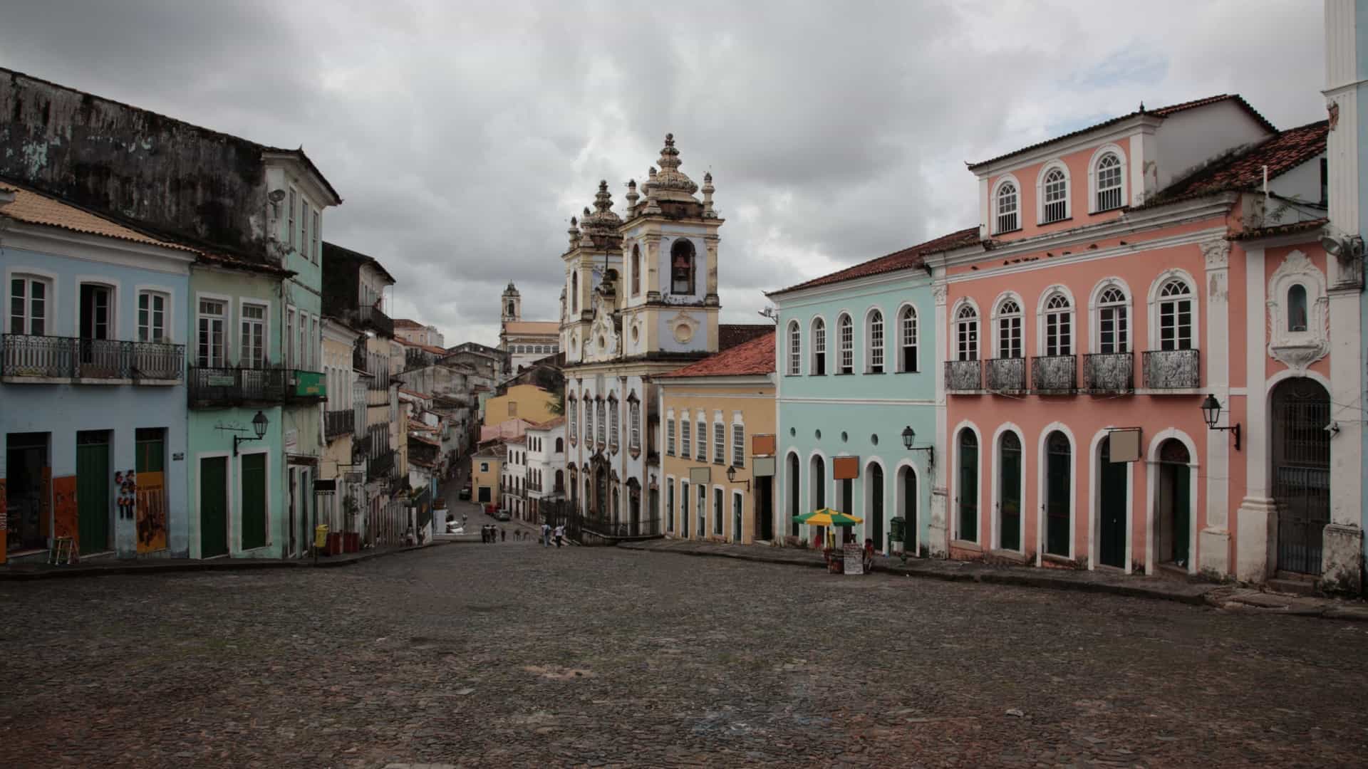 A dynamic, sunlit view of the UNESCO World Heritage Site of Pelourinho in Salvador, Bahia, Brazil. The cobblestone street is lined with beautifully restored, brightly painted colonial buildings featuring Portuguese Baroque architecture, reflecting the area's rich Afro-Brazilian culture and history.