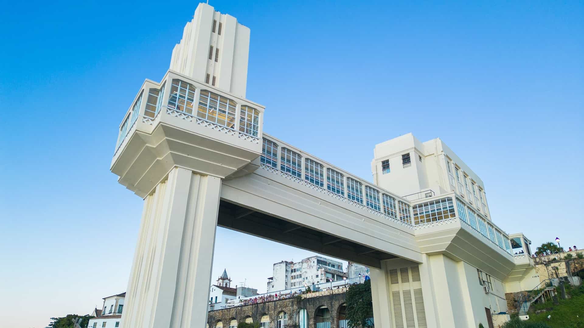 A wide panoramic view of the historic Salvador skyline and the Bay of All Saints, dominated by the towering, white, Art Deco structure of the Elevador Lacerda. This famous 72-meter public elevator connects the Upper City (Cidade Alta) to the Lower City (Cidade Baixa), offering a dramatic contrast between the modern architecture and the colonial buildings along the waterfront.