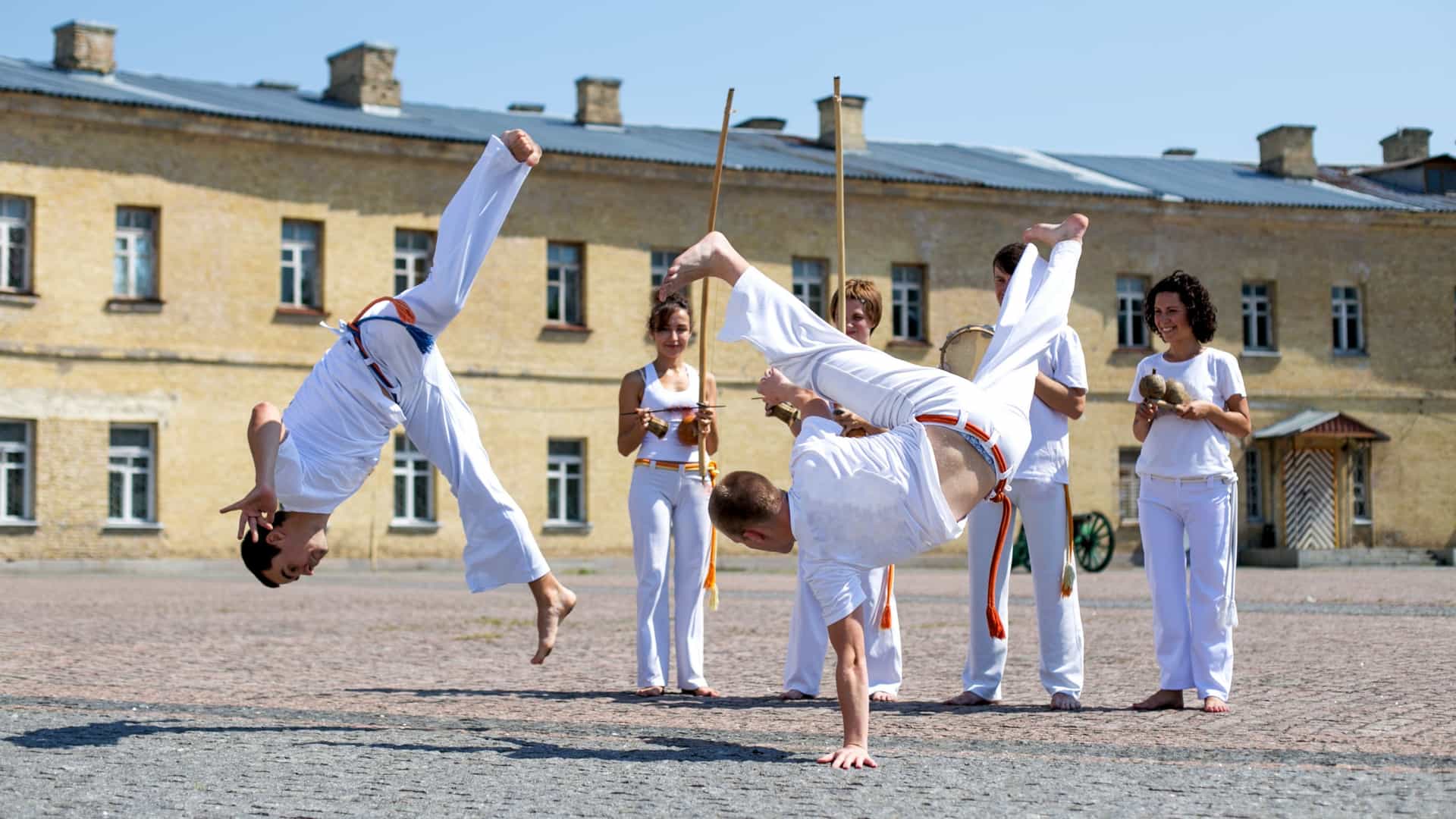 A dynamic outdoor demonstration of Capoeira, an Afro-Brazilian martial art. Two men in traditional white uniforms perform complex acrobatic kicks and handstands, while a group of people in the background plays traditional Capoeira instruments, including the berimbau.