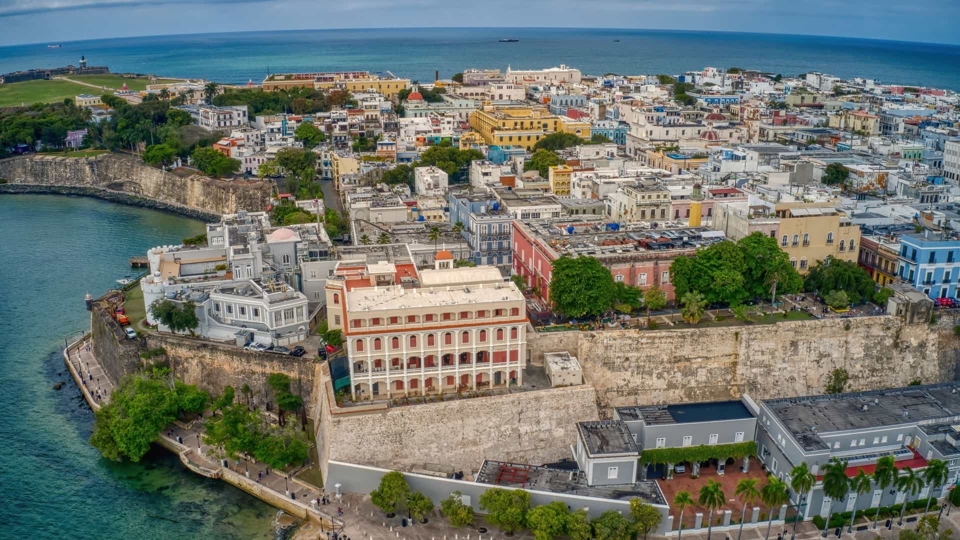  An aerial view of Old San Juan, Puerto Rico, showing the densely packed neighborhood of colorful colonial buildings with red tiled rooftops, which contrasts with the sprawling green lawn of the historic Castillo San Felipe del Morro fort in the background, all set against the blue waters of the ocean and the Atlantic sky.