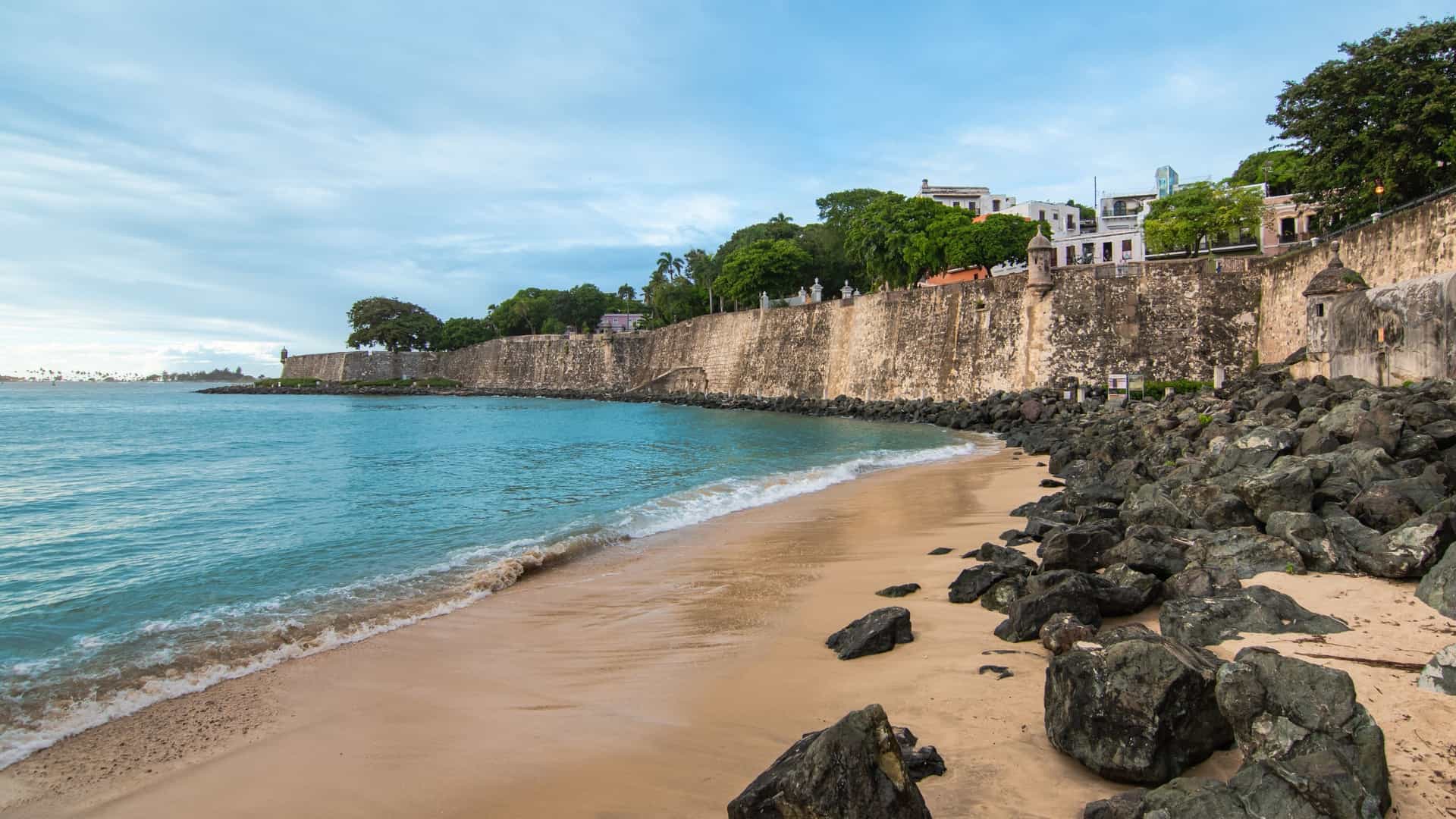  A scenic view of the historic stone city wall of Old San Juan, Puerto Rico, rising dramatically along the coastline of the Atlantic Ocean. Waves crash against the rocky base of the wall, and a small, stone sentry box (garita) is visible atop the wall in the distance, a characteristic feature of the city's Spanish colonial fortifications.