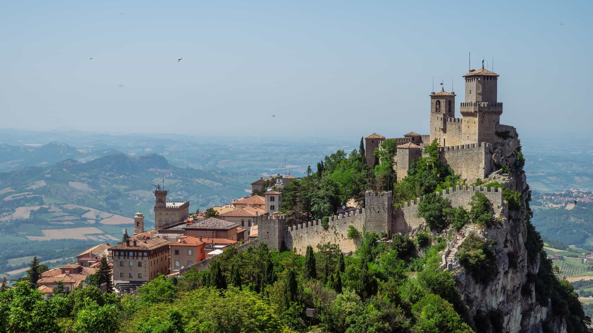  A majestic aerial view of the Three Towers of San Marino on the peaks of Monte Titano. The ancient stone fortresses, symbols of the Republic, stand proudly against a clear sky, surrounded by the lush green mountain slopes and the cityscape of San Marino below.