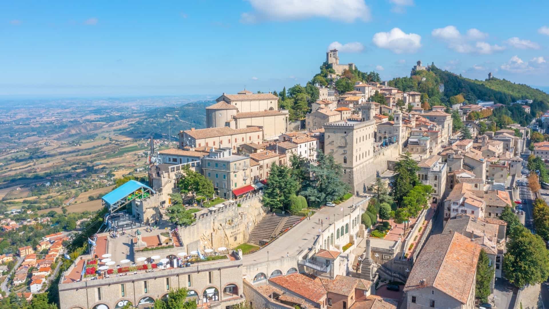  An aerial panoramic view of the historic Old Town of San Marino (Città di San Marino), perched on the steep ridge of Monte Titano. The image shows a dense cluster of medieval stone buildings with red tiled roofs and ancient fortification walls, with the surrounding terrain of lush green, rolling hills extending into the distance.