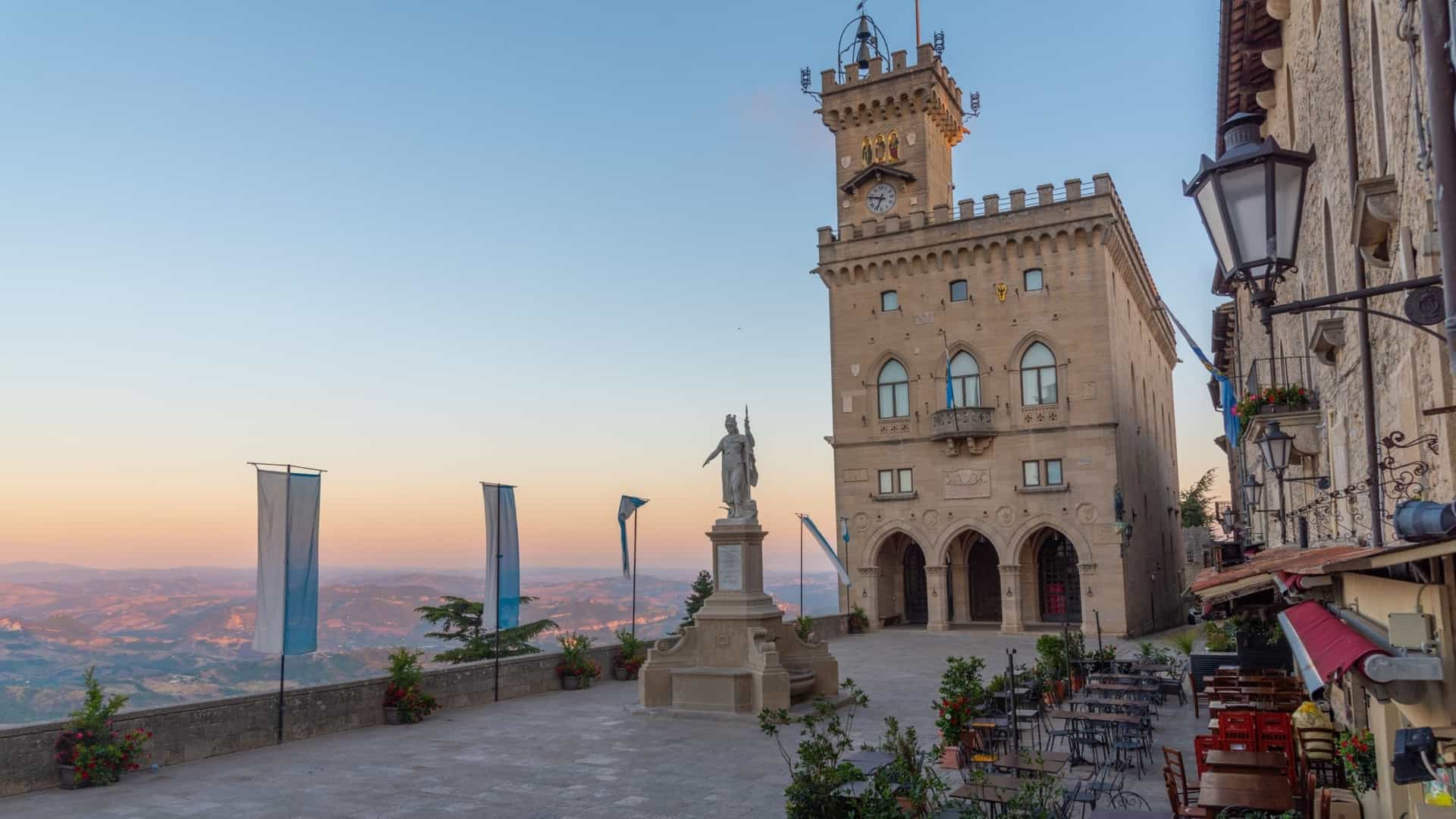  A sunrise view of the Palazzo Pubblico (Public Palace), the seat of government and town hall of the City of San Marino. The neo Gothic palace, with its distinctive crenellated tower and facade, is illuminated by the warm, golden light of dawn. It stands prominently in the Piazza della Libertà (Liberty Square), with the surrounding hills and cityscape visible in the background under a colorful sunrise sky.