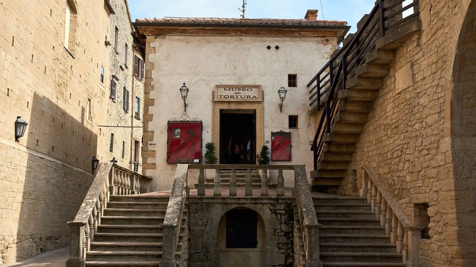  A dimly lit indoor exhibit within the Museum of Torture in San Marino. The image focuses on a collection of medieval torture instruments, with one prominent device resembling an Iron Maiden—a large vertical cabinet or sarcophagus with spikes on the inside. The instruments are displayed against rough stone walls, enhancing the chilling and historic atmosphere of the museum.