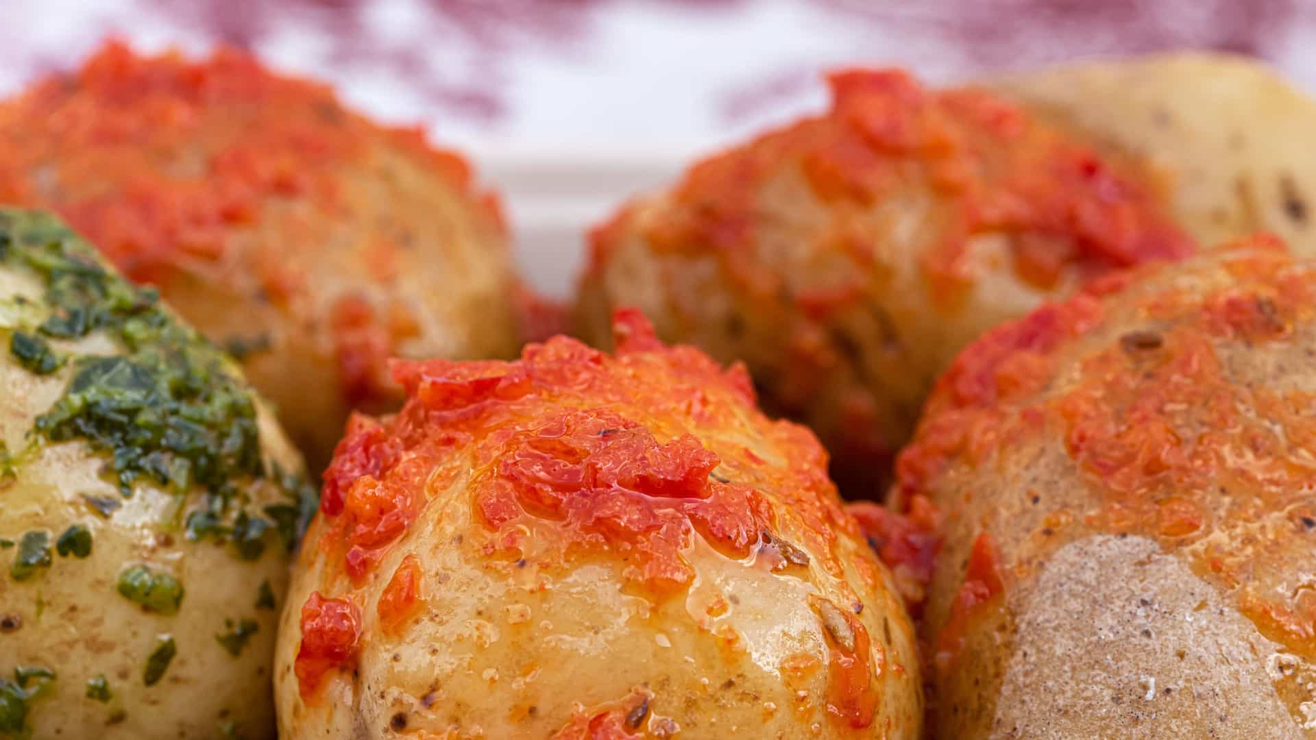  A close up of a serving of Papas Arrugadas, the typical Canarian dish of small, unpeeled potatoes boiled in saltwater until their skins are wrinkled and coated with a fine white salt crust. The potatoes are arranged on a white plate with two traditional dipping sauces on the side: Mojo Rojo (a red, spicy sauce made with chili peppers and paprika) and Mojo Verde (a green sauce made with cilantro or parsley).