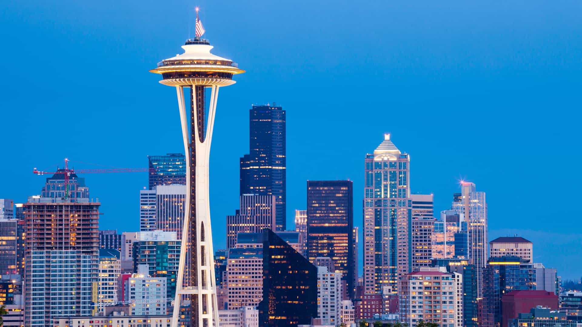 A dusk or night-time photo of the Seattle skyline. The Space Needle dominates the foreground, lit up against a deep blue sky. Behind it, a cluster of downtown skyscrapers are visible, with windows illuminated. Trees are visible in the lower foreground.