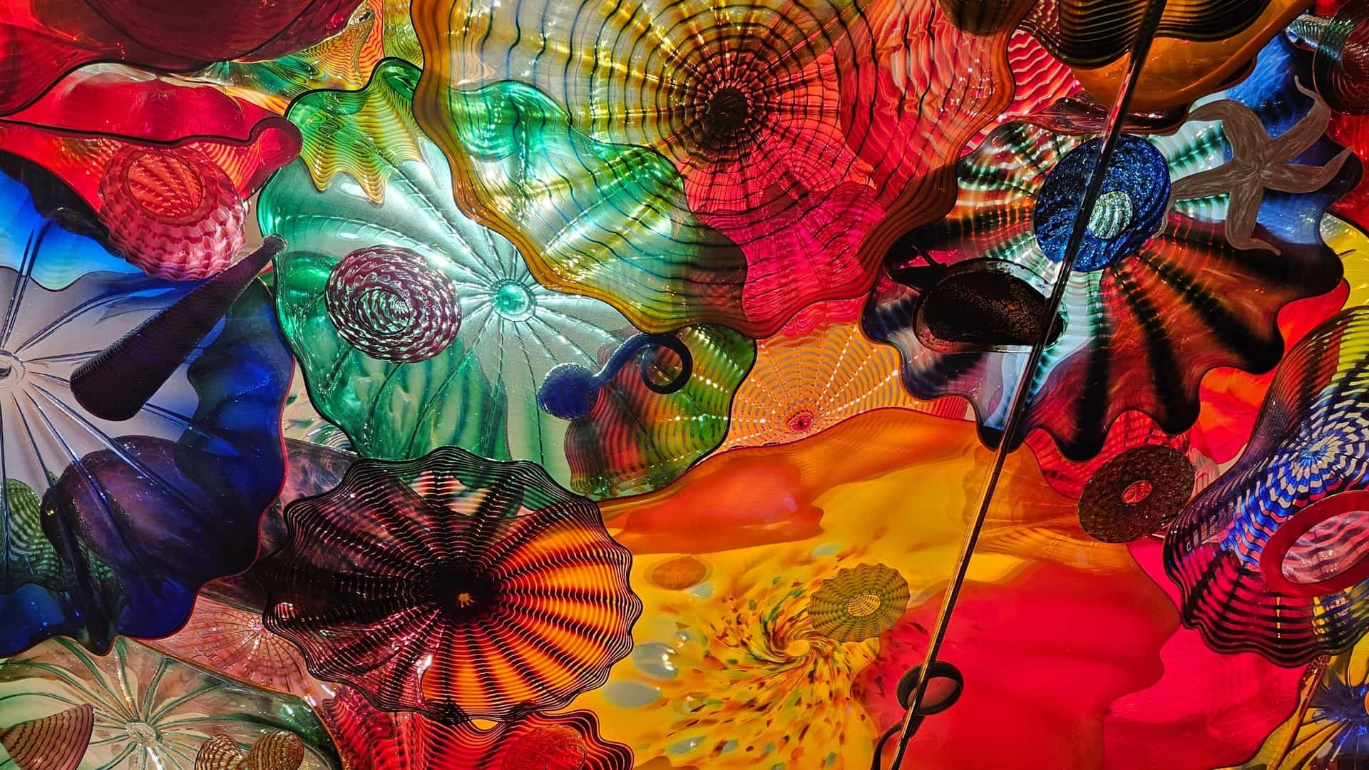 A vibrant interior view of the Chihuly Garden and Glass exhibition in Seattle, showcasing a massive, multi-colored glass sculpture by artist Dale Chihuly. The ceiling is adorned with numerous brilliant glass pieces, likely part of a Chandelier or the Persian Ceiling exhibit, displaying intricate forms and a dazzling array of colors, emphasizing the artist's use of light and glass art.