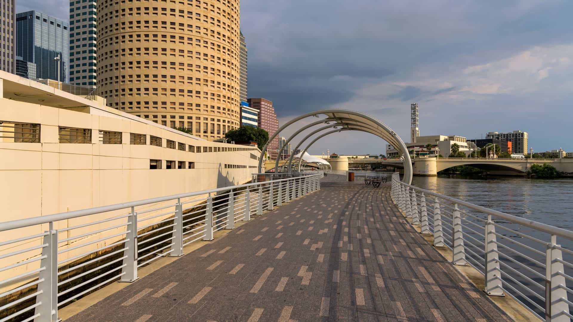A wide-angle view of the Tampa Riverwalk in Florida, showing the pedestrian path winding along the Hillsborough River. The distinct Moorish architecture of the University of Tampa (former Tampa Bay Hotel) and the Kennedy Boulevard Bridge are visible in the background on a calm evening.