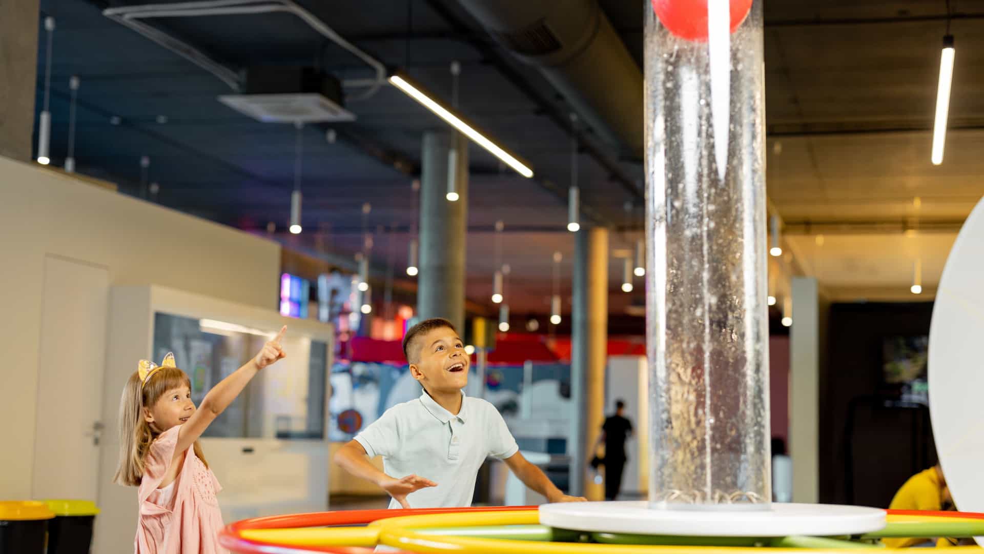 A young boy and girl are excitedly watching a red ball float in a tall, clear vertical tube of air or water, which is part of a physics or air-pressure exhibit at the Museum of Science & Industry (MOSI) in Tampa. The boy has his hands raised, and the girl is pointing toward the ball.