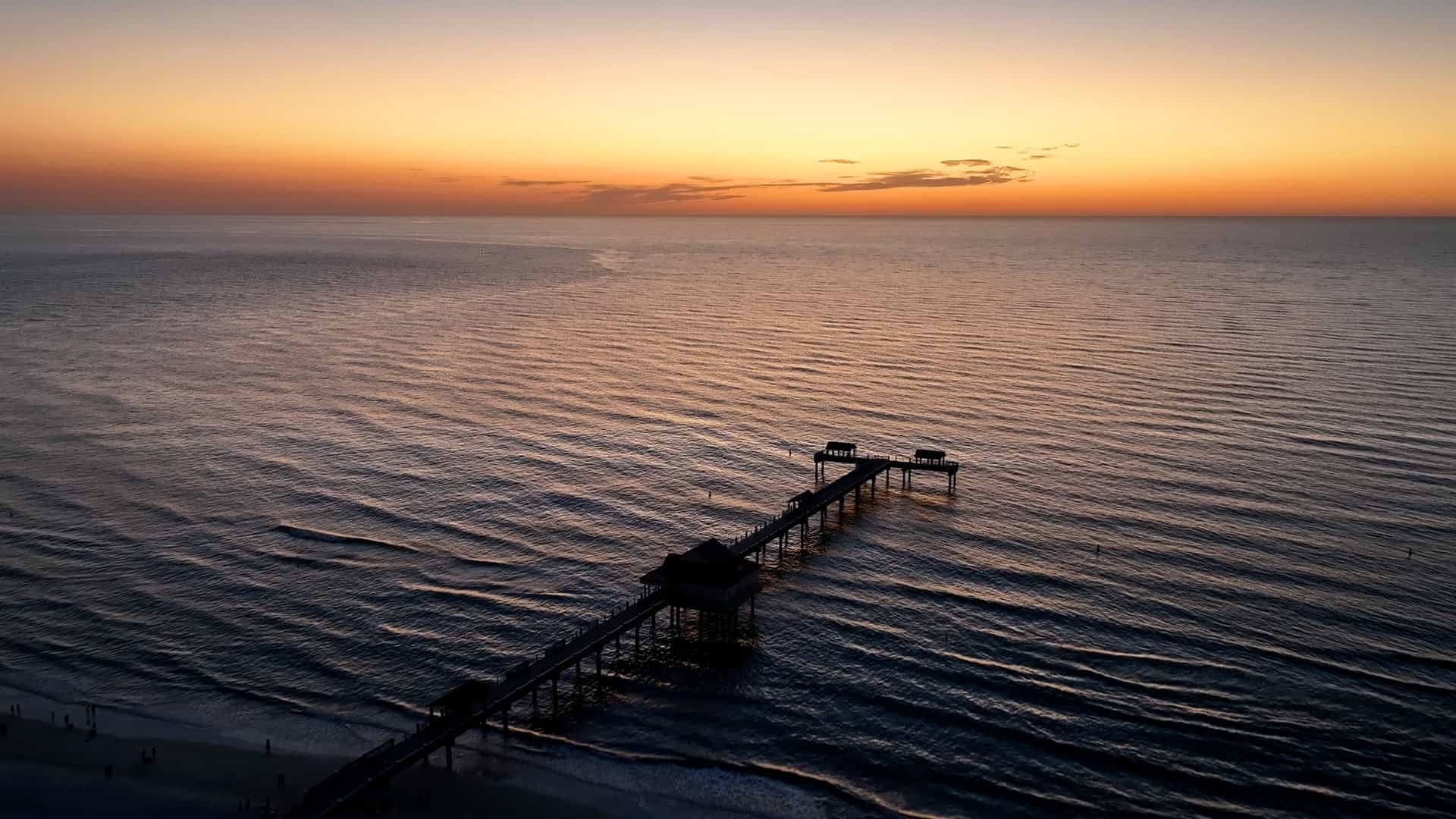 A beautiful sunset at Pier 60, Clearwater Beach, Florida, United States. The long, concrete fishing pier extends into the Gulf of Mexico, with the structure silhouetted against a brilliantly colored sky of orange, red, and pink hues. The Gulf's water reflects the vibrant colors of the sunset.