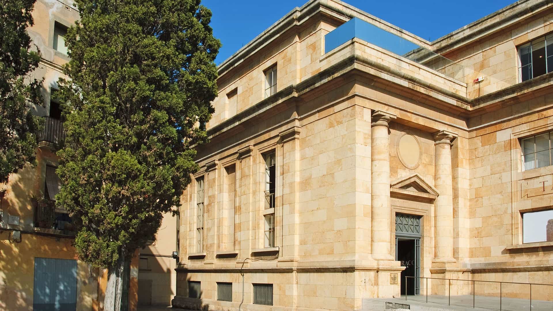  The corner and main entrance of a large, stately building with a facade of yellow beige stone, featuring classical architectural elements like columns and cornices, with tall, narrow windows. A dark green cypress tree stands prominently to the left, casting a shadow on the building under a clear blue sky. This is the Museu Nacional Arqueològic de Tarragona (MNAT) in Spain.