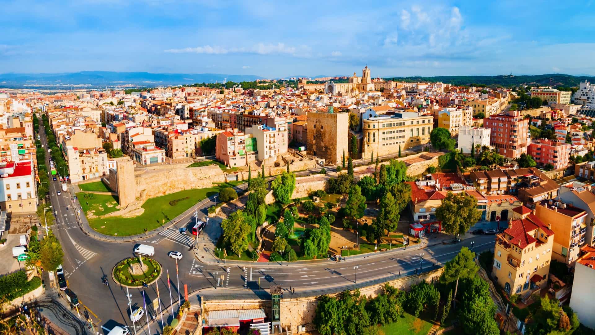  A panoramic aerial view of the city of Tarragona, Spain, showing the extensive port area with ships docked, the dense layout of the city's buildings and terracotta rooftops, and the coastline meeting the deep blue of the Mediterranean Sea.