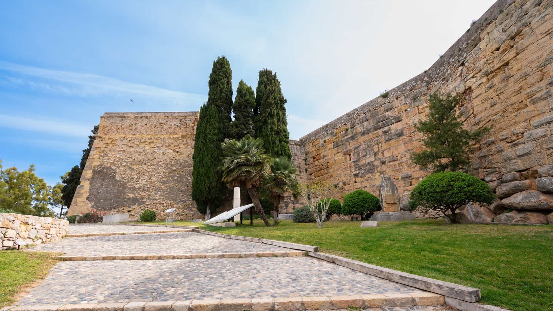  A view along the Passeig Arqueològic (Archaeological Walk) in Tarragona, Spain, showing the massive, ancient Roman walls constructed of large stone blocks. The pathway runs alongside the monumental walls, which are topped by battlements, and is lined with trees and greenery.