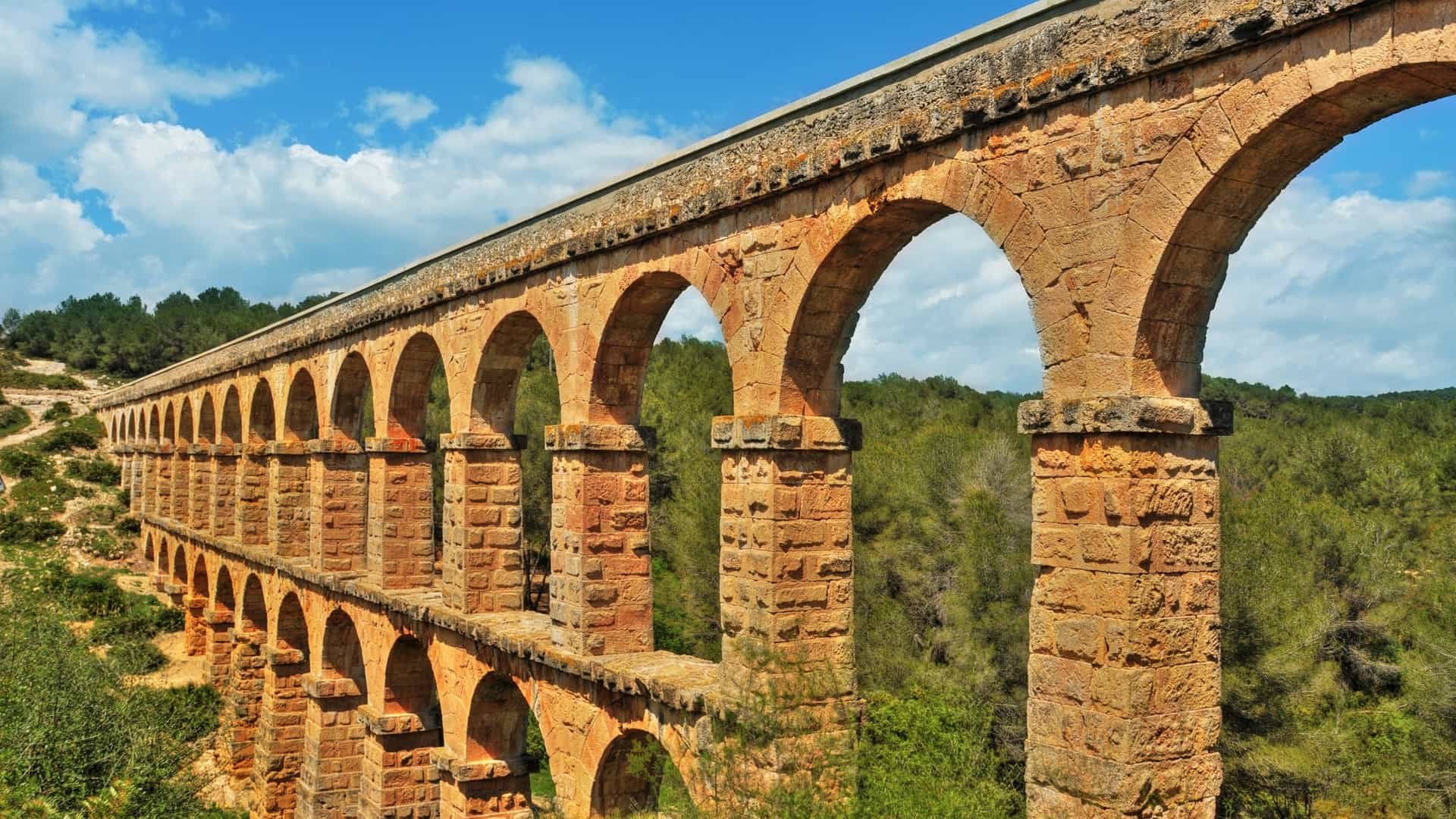  A view of the Pont del Diable (Devil's Bridge), a monumental Roman aqueduct near Tarragona, Spain, rising from a densely wooded area. The aqueduct consists of two tiers of arches—eleven on the lower level and twenty five on the upper—built of large, dry laid stone blocks, and is seen crossing a small valley.