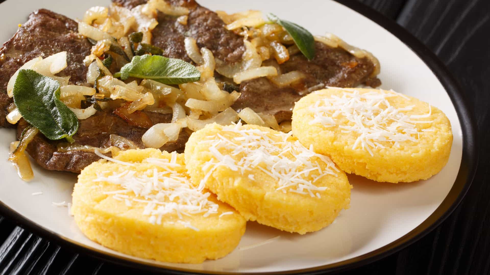  A dish of Fegato alla Veneziana (Venetian style liver and onions) is presented in a white, shallow bowl. Thin slices of tender calf's liver, which are dark brown, are combined with a generous portion of caramelized, translucent white onions. The dish is garnished with fresh, chopped sage leaves and is likely prepared with a splash of vinegar or white wine for a sweet and sour flavor balance.