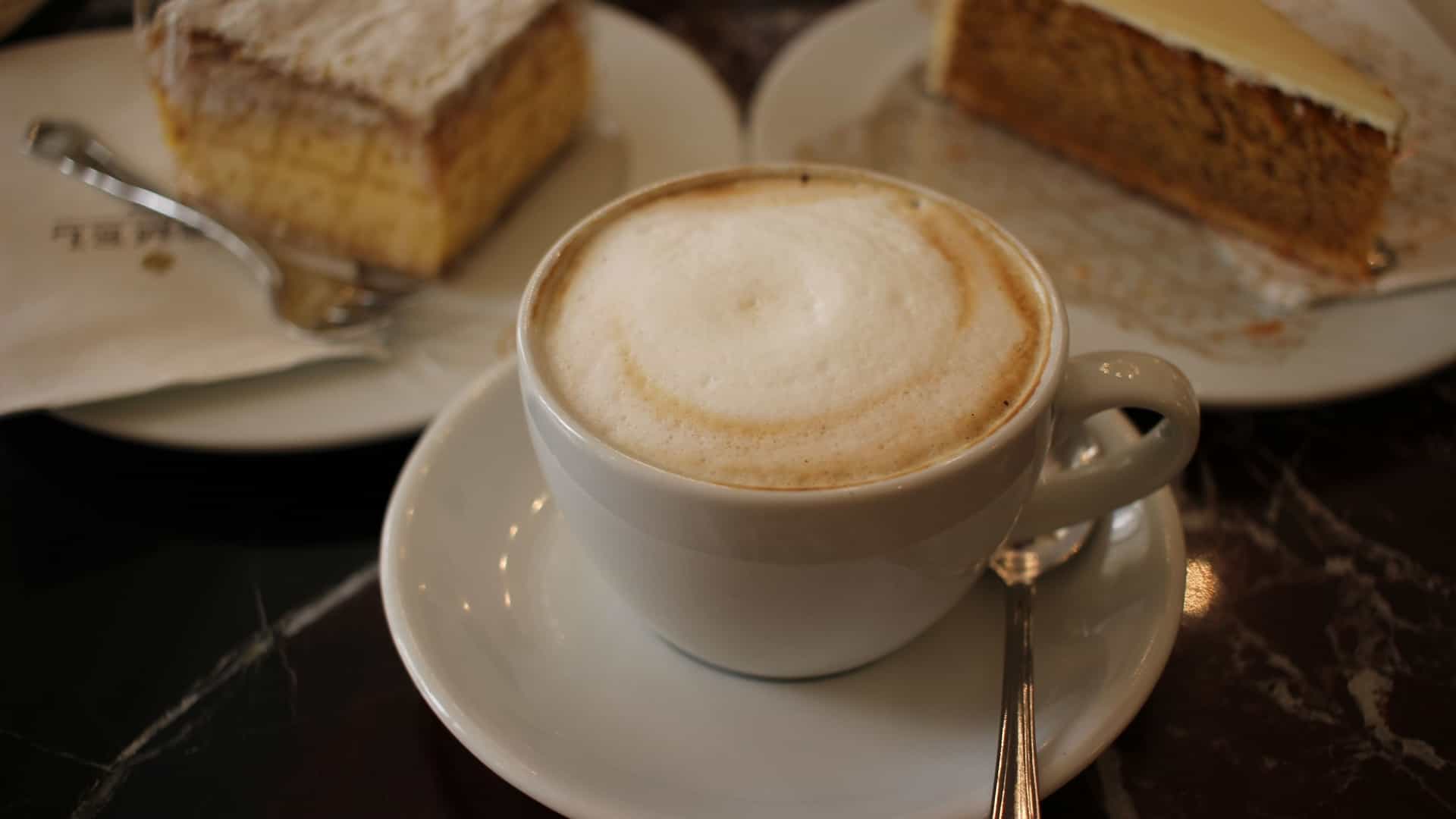  A "Wiener Melange," a classic Viennese coffee specialty similar to a cappuccino, is served in a white ceramic cup on a matching square saucer. The cup is filled with coffee, steamed milk, and a layer of milk foam on top, often sprinkled with a small amount of cocoa or cinnamon. A metal spoon rests on the saucer next to the cup.