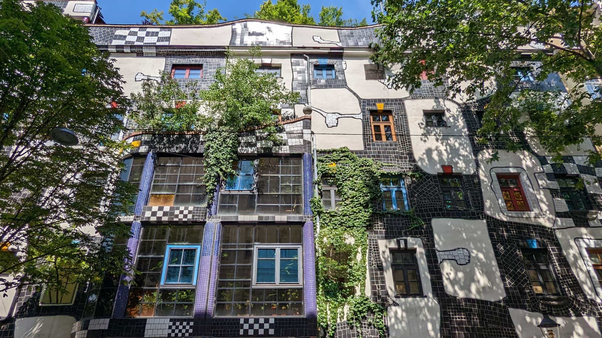 The vibrant, asymmetrical exterior facade of the Kunst Haus Wien (Hundertwasser Museum) in Vienna, which features an irregular, checkerboard pattern of bright, mismatched colors, a notable lack of straight lines, and foliage growing out of the building.