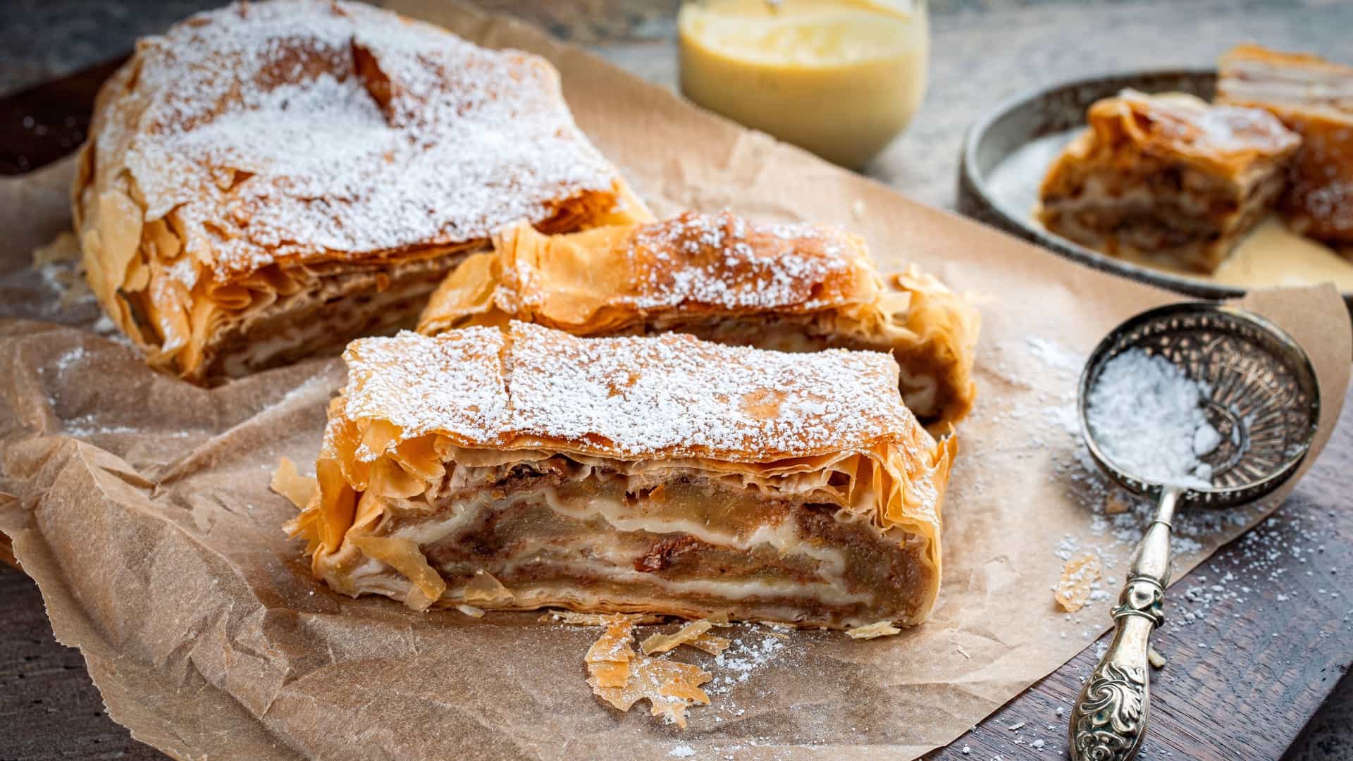  A close up view of a slice of Traditional Bavarian Apfelstrudel with a flaky crust, generously filled with baked apples and raisins, dusted with powdered sugar, and served with a side of vanilla sauce in a small white dish.