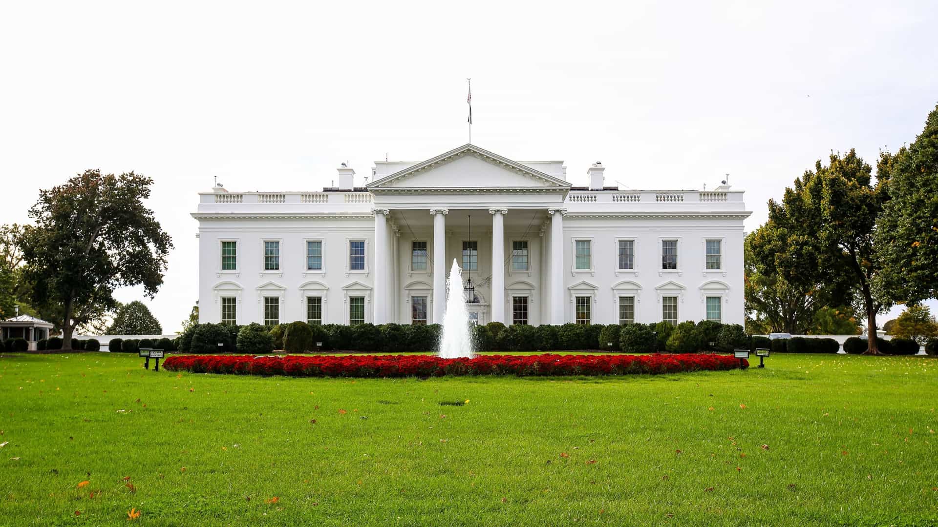  The North Portico of the White House, the official residence of the U.S. President, is viewed from Pennsylvania Avenue in Washington, D.C. The neoclassical mansion features a prominent columned portico and is surrounded by green lawn and trees, under a cloudy blue sky.