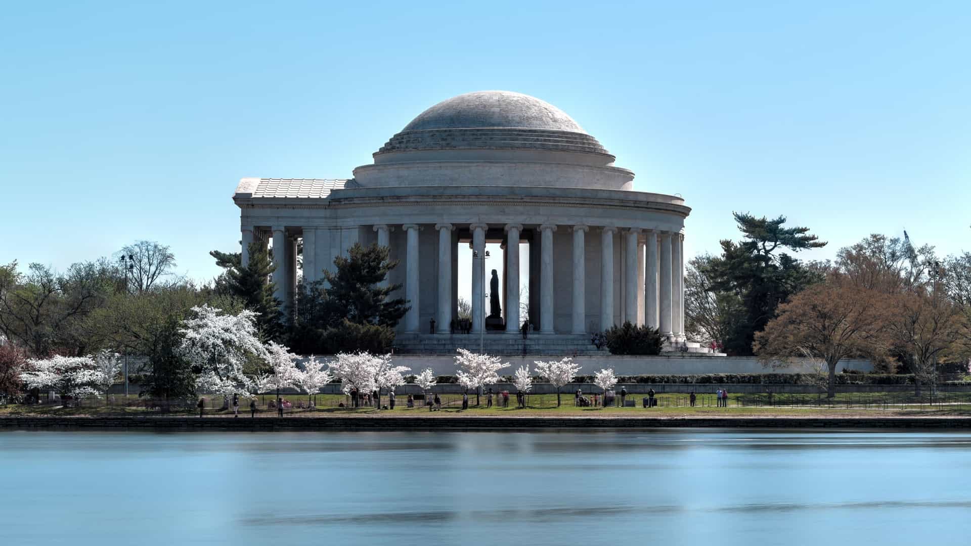  The Thomas Jefferson Memorial in Washington, D.C., a white domed, neoclassical rotunda, is seen across the Tidal Basin. The memorial is framed by a dense canopy of pink and white cherry blossoms during the annual Cherry Blossom Festival, with the monument's reflection visible in the water.