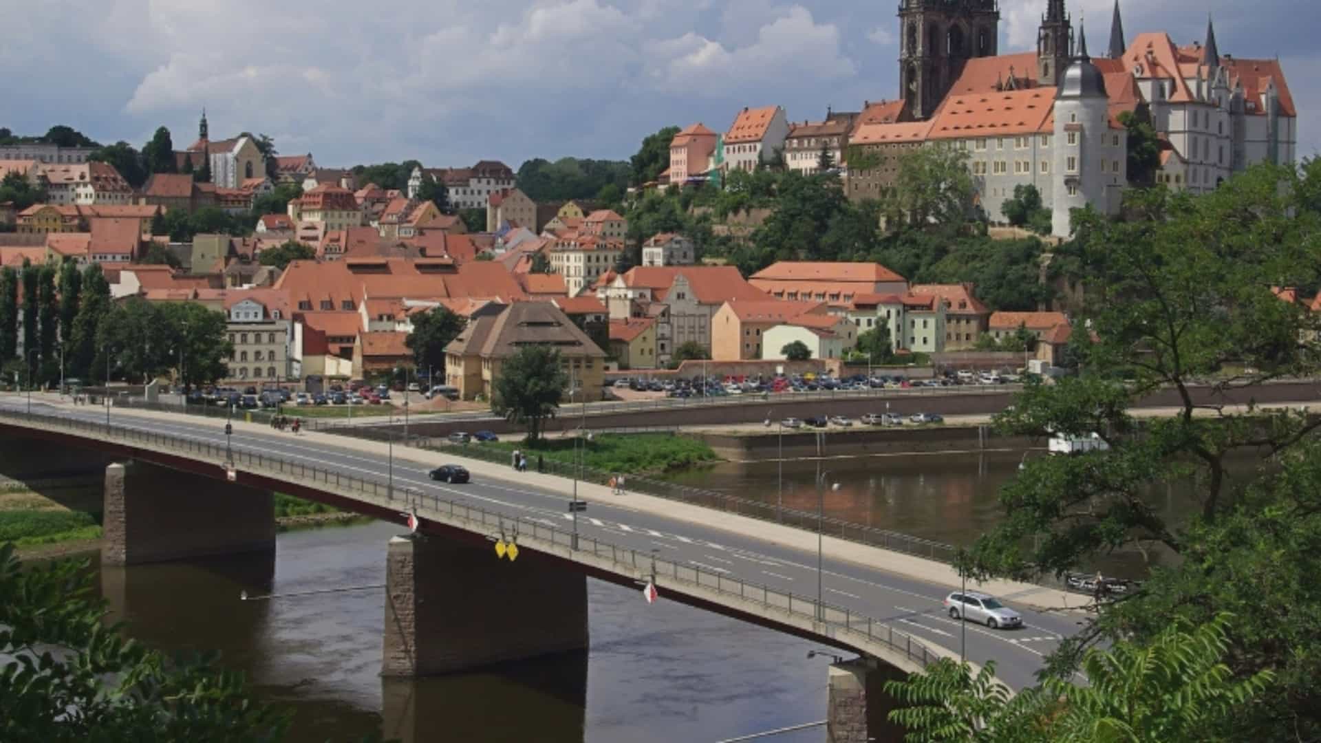 An elevated view of the historic city of Meissen, Germany, located on the Elbe River. The image shows a bridge crossing the river in the foreground, with traffic passing over it. The cityscape rises on the opposite bank, with a cluster of buildings with red-tiled roofs. Perched on a hill above the city is the prominent Albrechtsburg Castle and the Meissen Cathedral with its two tall, gothic spires. The sky is partly cloudy, adding to the dramatic atmosphere of this historical Elbe River city.