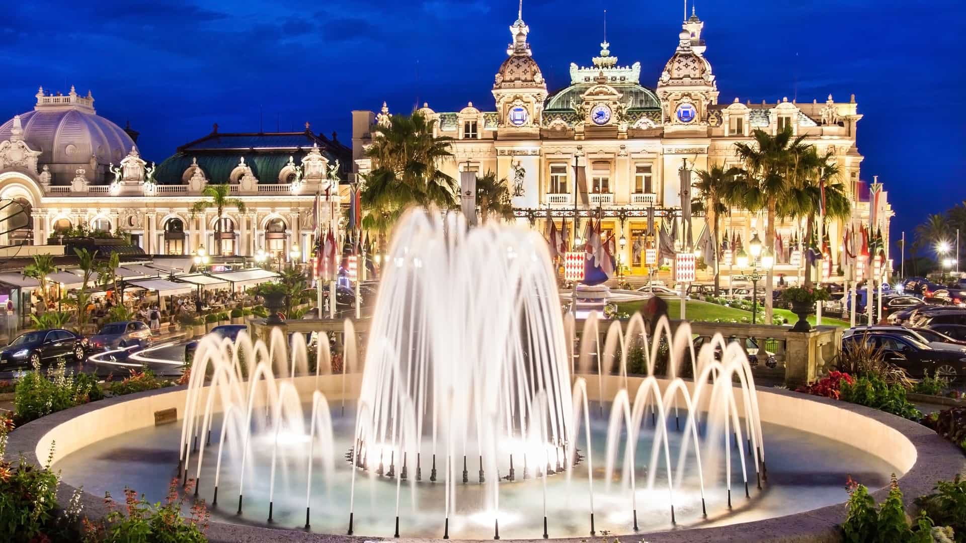 An elegant nighttime view of the Monte Carlo Casino in Monaco, a famous landmark in Europe. The magnificent building is brightly lit, showcasing its ornate Belle Époque architecture with grand domes and intricate details. In the foreground, a large, illuminated fountain sprays water high into the air. The surrounding area features palm trees, manicured gardens, and other illuminated buildings under a deep blue evening sky. The image captures the luxury and glamour of this iconic European city-state.