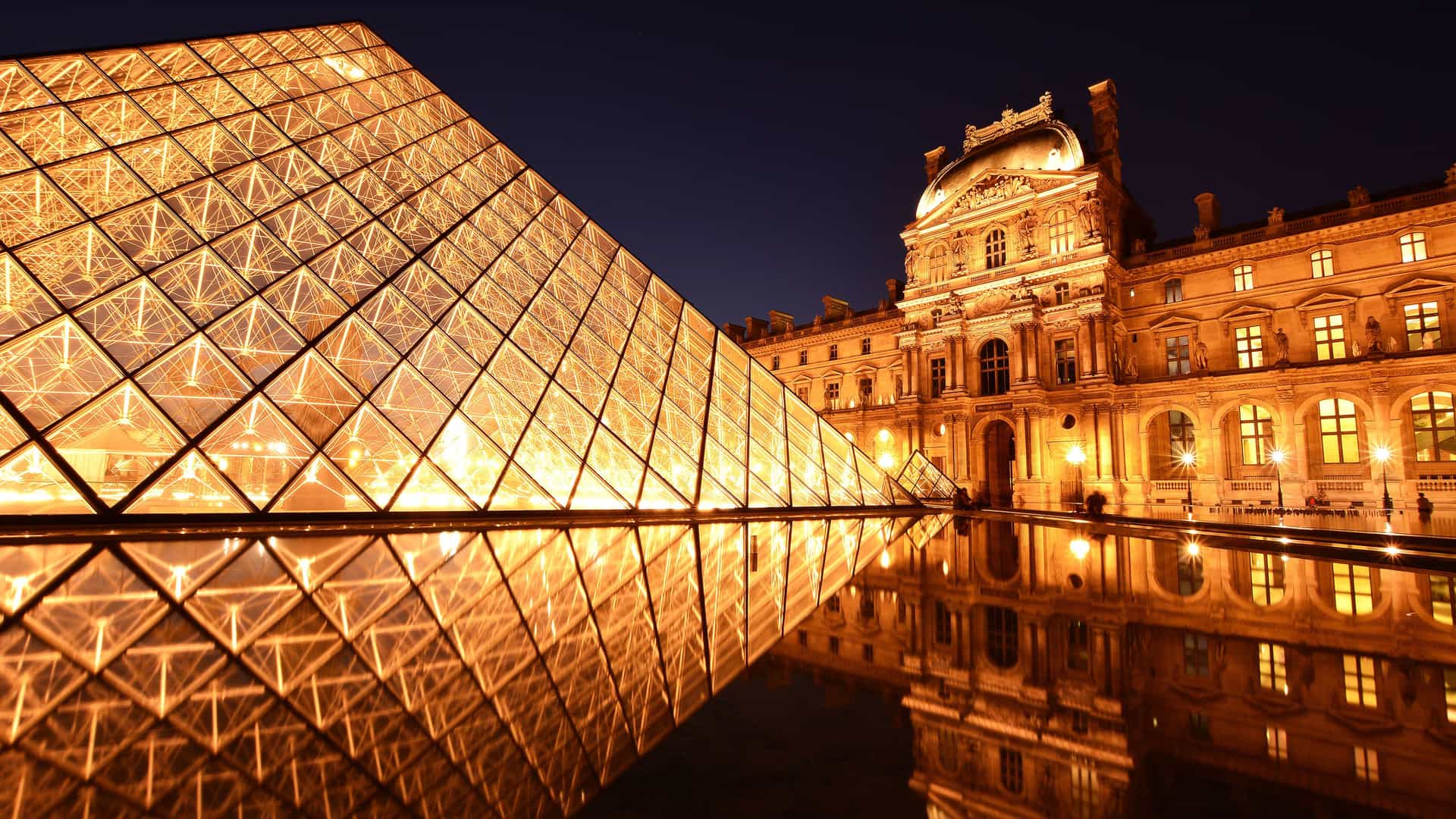 An elegant nighttime view of the Louvre Museum in Paris, France. The iconic glass pyramid entrance is brilliantly illuminated with a warm golden light, reflecting on the still water in the foreground. In the background, a section of the historic museum building is also lit up, showcasing its intricate architecture. The dark blue sky provides a striking contrast, highlighting this famous landmark of European art and culture.