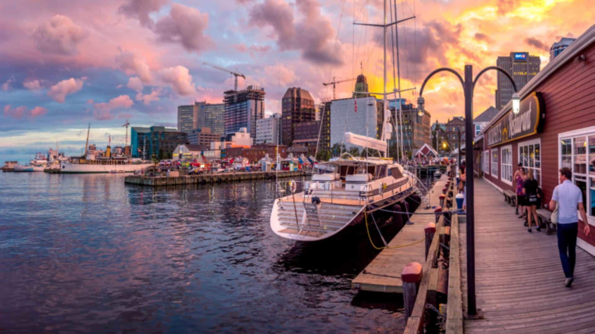 A wide-angle, sunset view of the waterfront in Halifax, Nova Scotia, with a large white yacht docked at a pier lined with people walking. In the background, modern city buildings are visible against a sky of vibrant pink, orange, and purple clouds.