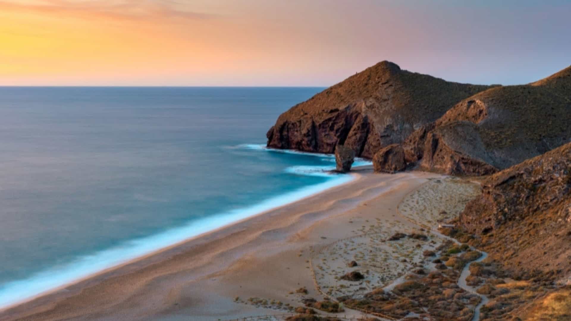An aerial view of the beautiful and secluded Playa de los Muertos in Puerto Vallarta, Mexico. The sandy beach is bordered by rugged, rocky cliffs and a serene blue ocean. The foamy white waves gently roll onto the shore, creating a soft, blurred effect. The sky is a gradient of soft orange and blue, indicating either a sunrise or sunset. The image captures the untouched and scenic coastline of this Mexican region.