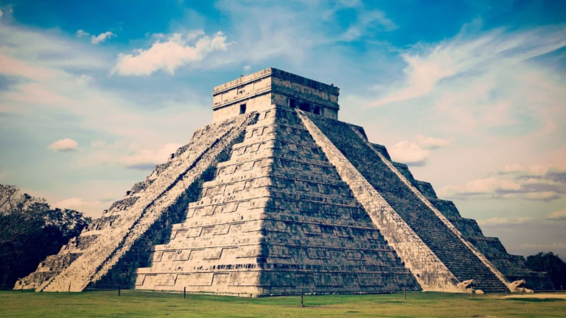 A striking view of the ancient Maya pyramid of El Castillo, or the Temple of Kukulcan, at Chichen Itza in the Yucatan region of Mexico. The large stepped pyramid stands on a green, grassy field under a brilliant blue sky with scattered white clouds. The sun casts a shadow on one side of the pyramid, highlighting the intricate stonework and the long staircases leading to the temple at the top. The image captures the grandeur and historical significance of this iconic Mexican landmark.