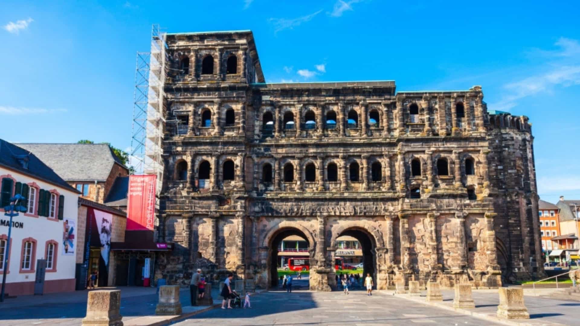 The ancient Roman gate, Porta Nigra, in the city of Trier, Germany. The large, dark gray stone structure has two arched passageways at its base and multiple tiers of arched windows above. Scaffolding is visible on the left side of the gate. In the foreground, a paved plaza with stone bollards is mostly empty, with a few people walking in the distance. The background shows parts of the city with historical buildings. The sky is a clear, bright blue, highlighting this famous landmark of the Moselle River region.