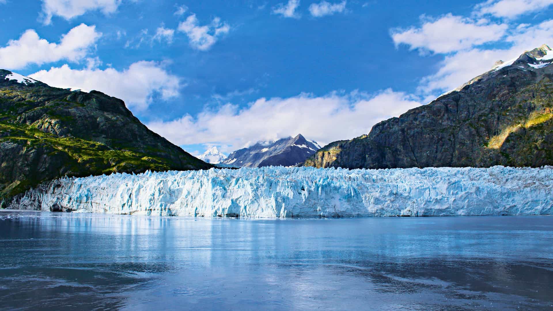 Norwegian Cruise Line ship sailing past a massive blue glacier in Glacier Bay, Alaska, with snow-capped mountains under a clear sky.