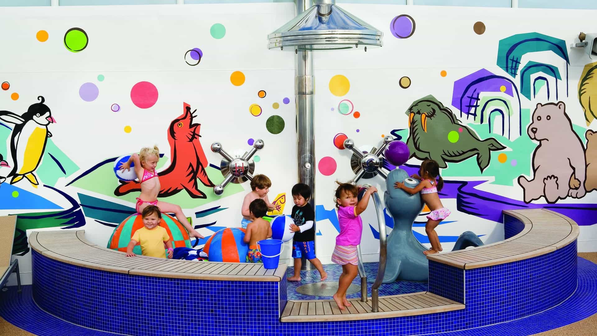 Children are playing at a colorful splash pad on the Norwegian Cruise Line's Norwegian Gem ship, with one child climbing on a gray seal statue and others sitting on the edge of the shallow pool.