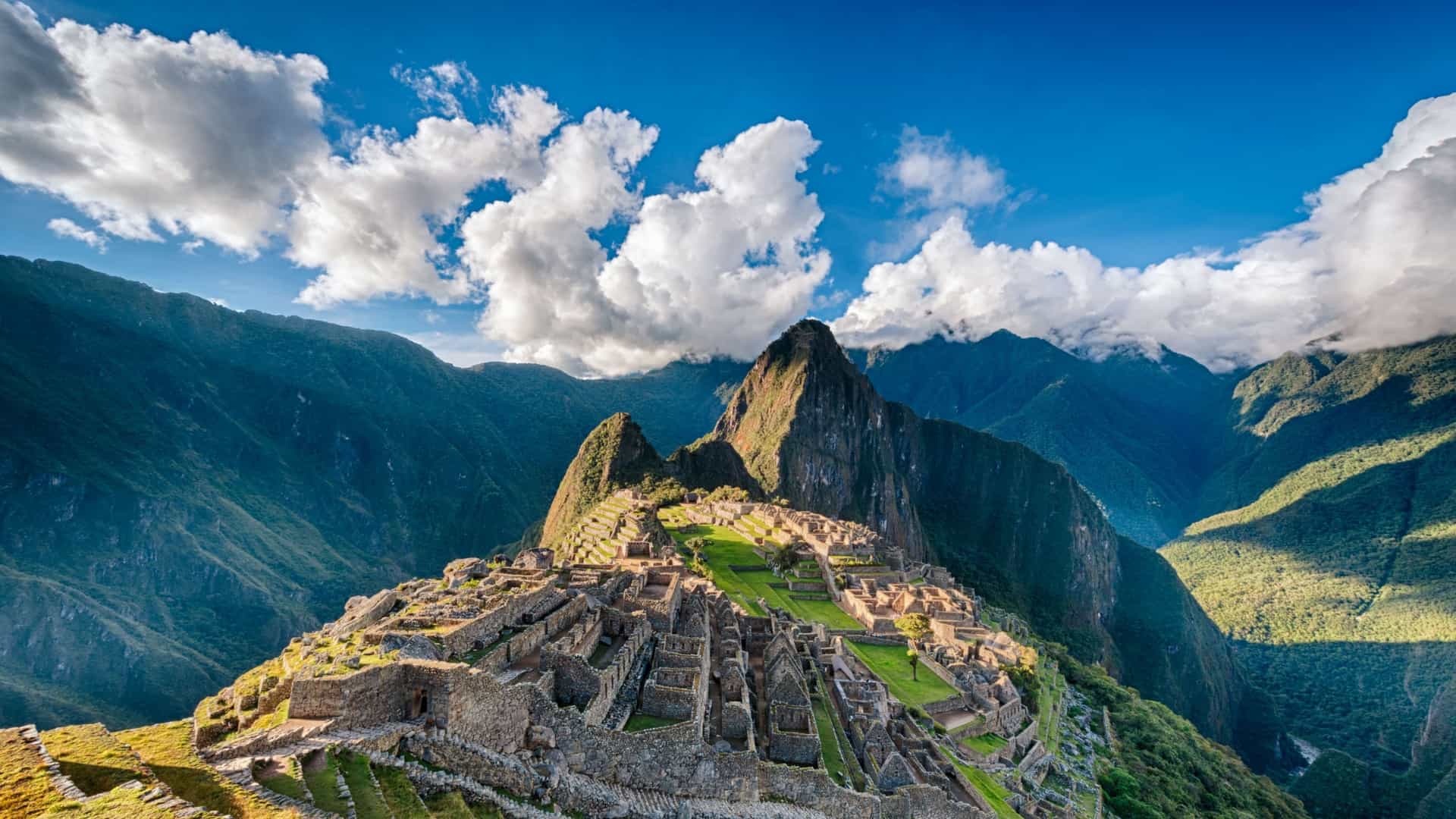 A breathtaking panoramic view of the ancient Inca citadel of Machu Picchu, nestled high in the Andes Mountains with dramatic peaks and a partly cloudy blue sky.