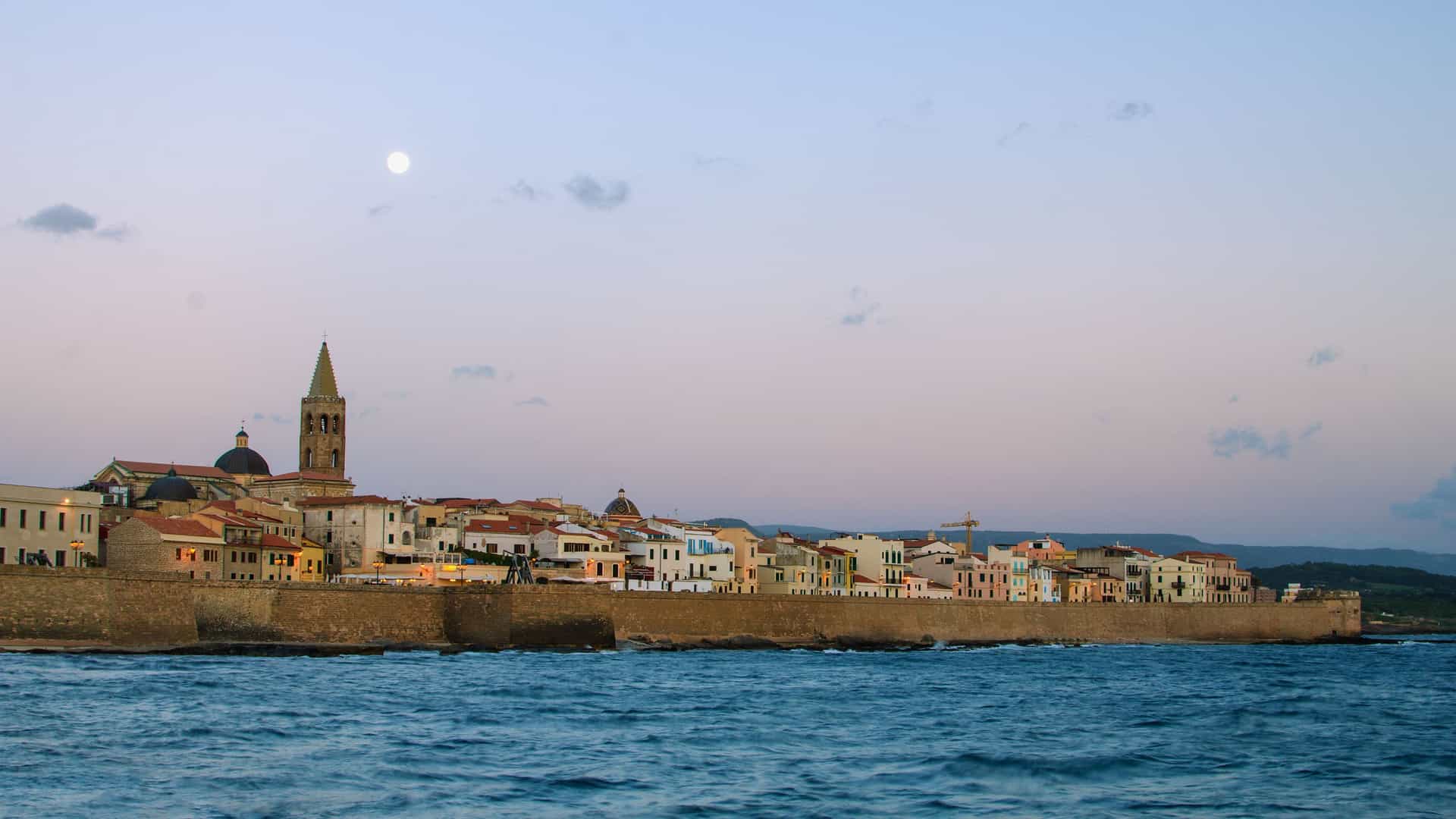 A panoramic twilight shot of Alghero, Italy, with its historic city walls and a bell tower silhouetted against a pastel sky with a full moon, reflecting on the Ligurian Sea.