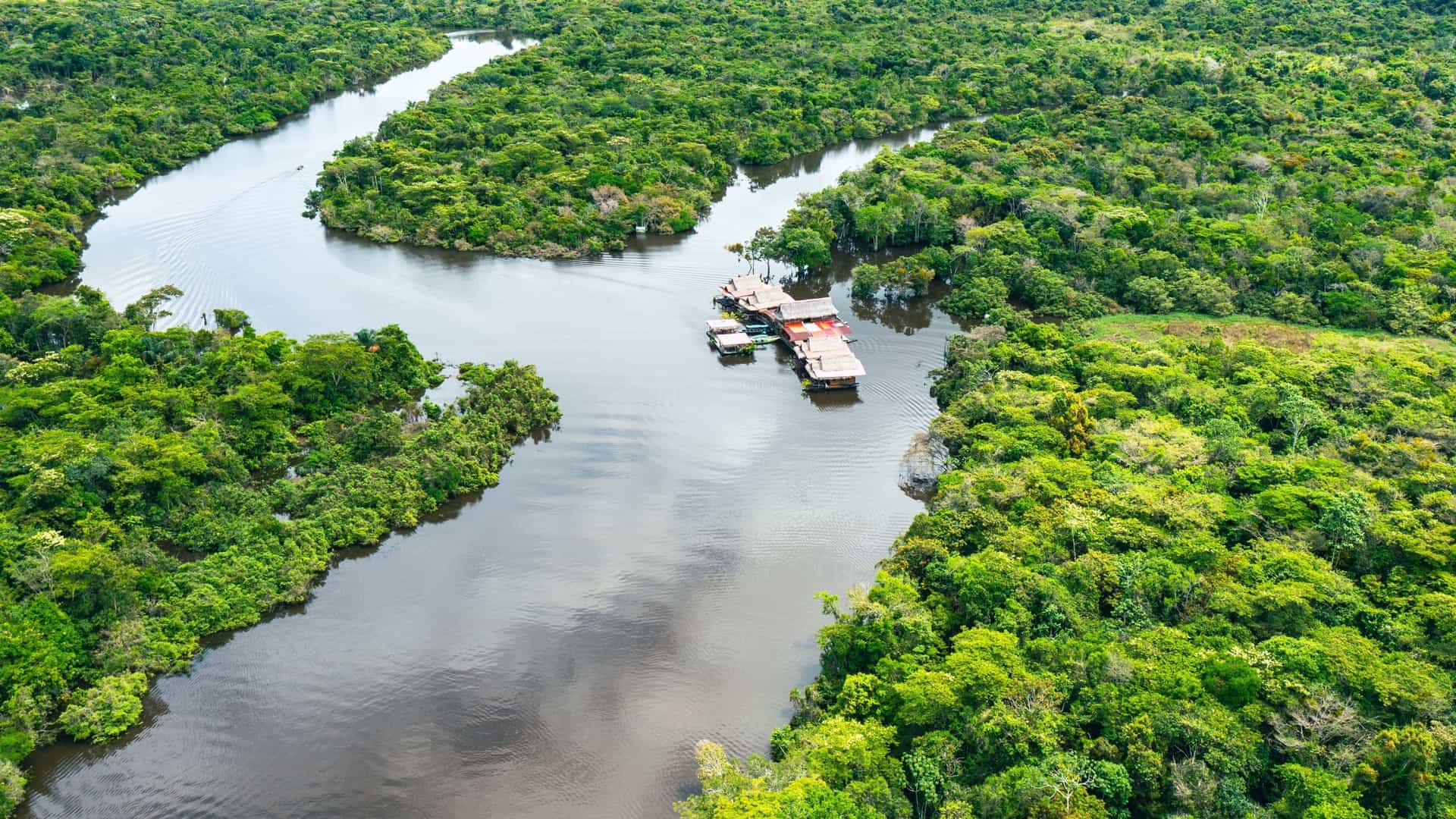 An aerial view of the vast Amazon River in Peru, meandering through a dense, green rainforest with a small floating lodge anchored along its banks.