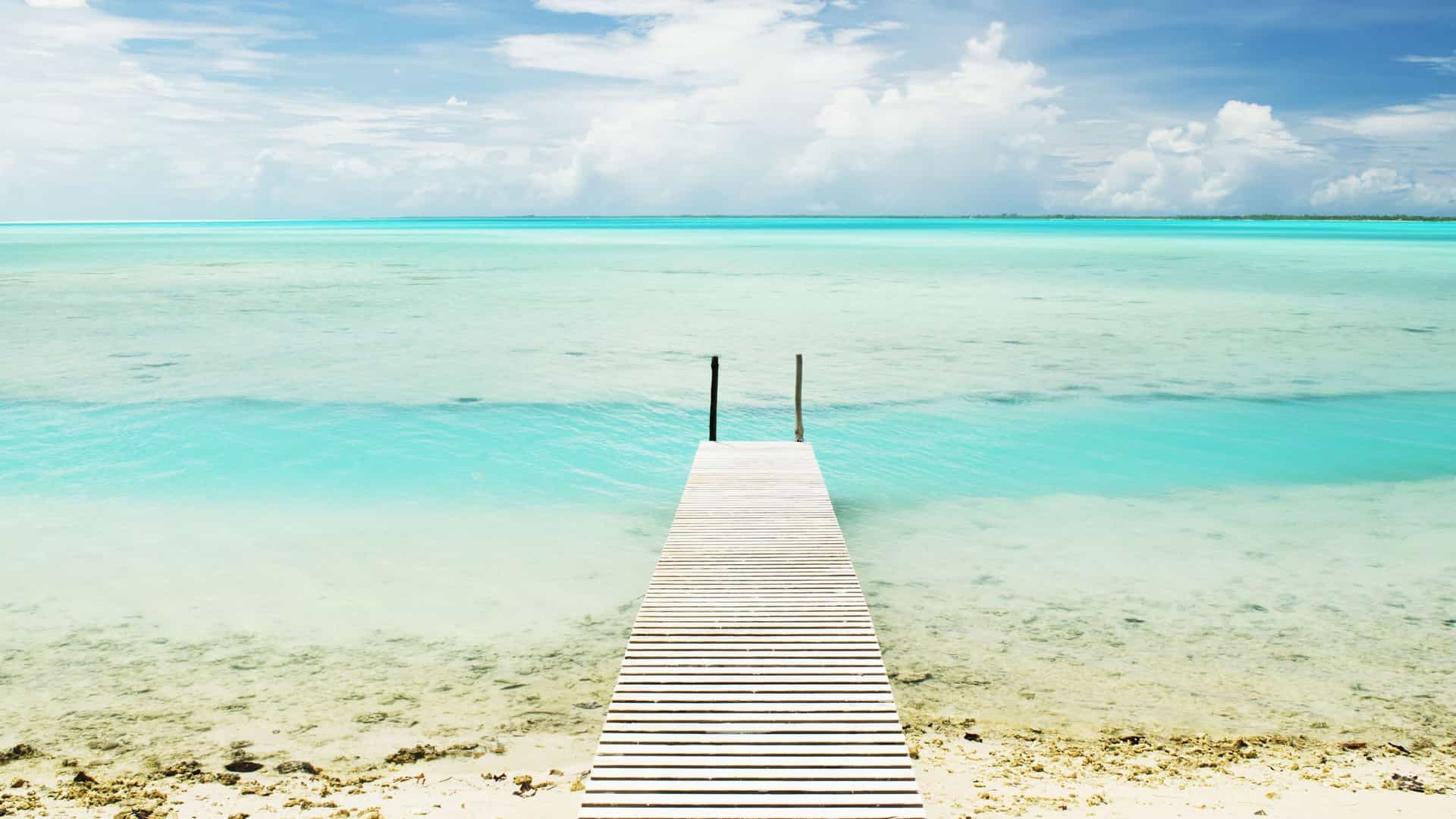 A scenic view of a wooden pier stretching out over the shallow, turquoise waters of a lagoon on Anaa atoll in French Polynesia, with a white sand beach.