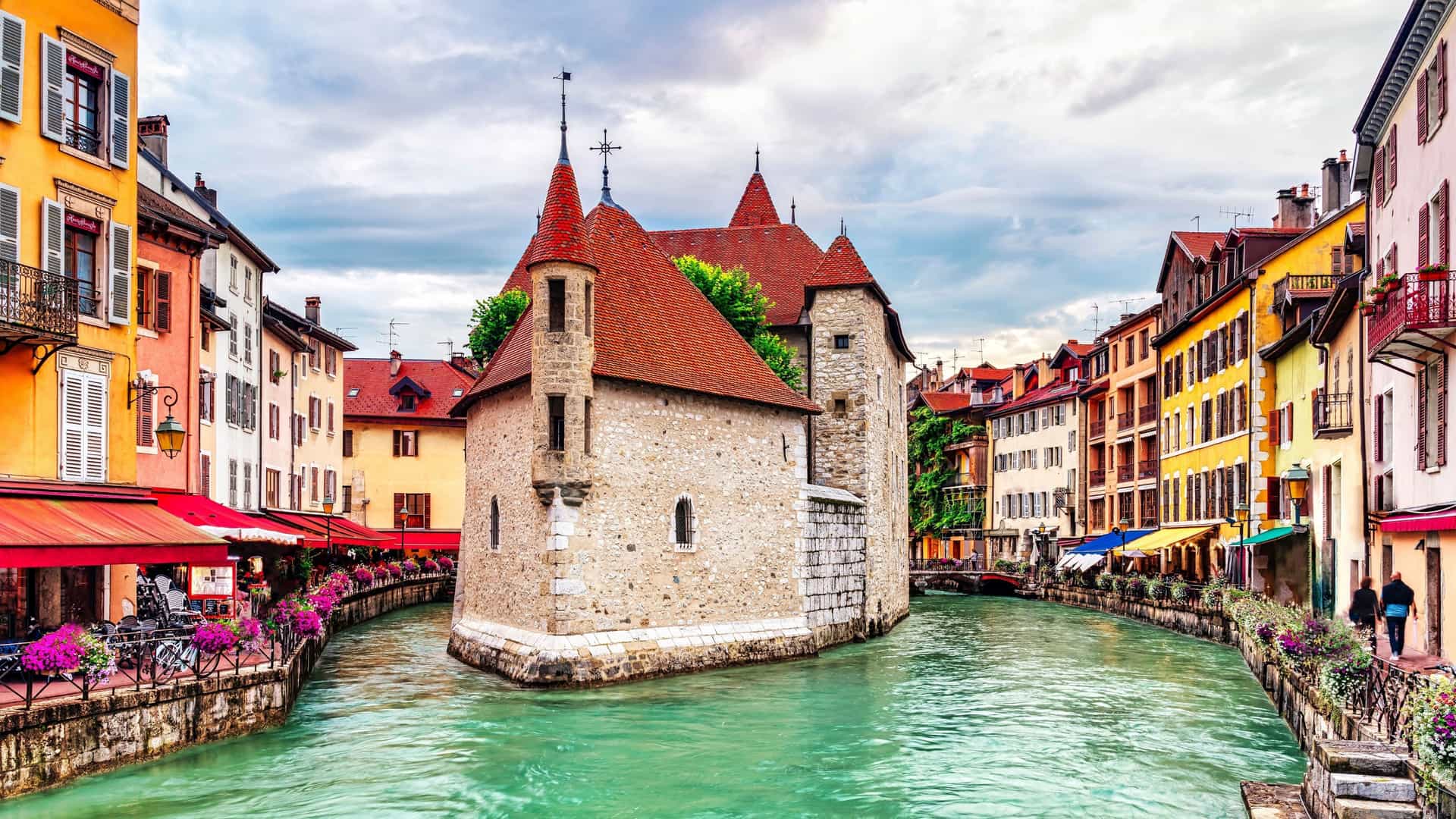 A vibrant scenic view of the medieval Palais de l'Île, a castle-like building in Annecy, France, situated in the middle of a canal with colorful buildings lining the banks.