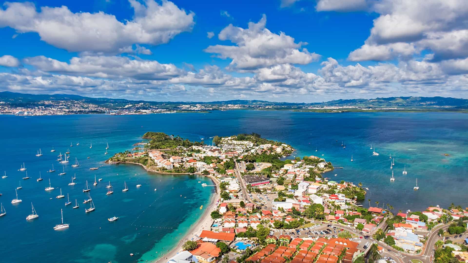 An aerial view of Anse Mitan in Martinique, a beautiful Caribbean village with a bustling beach, sailboats moored in the bay, and lush green hills in the distance.