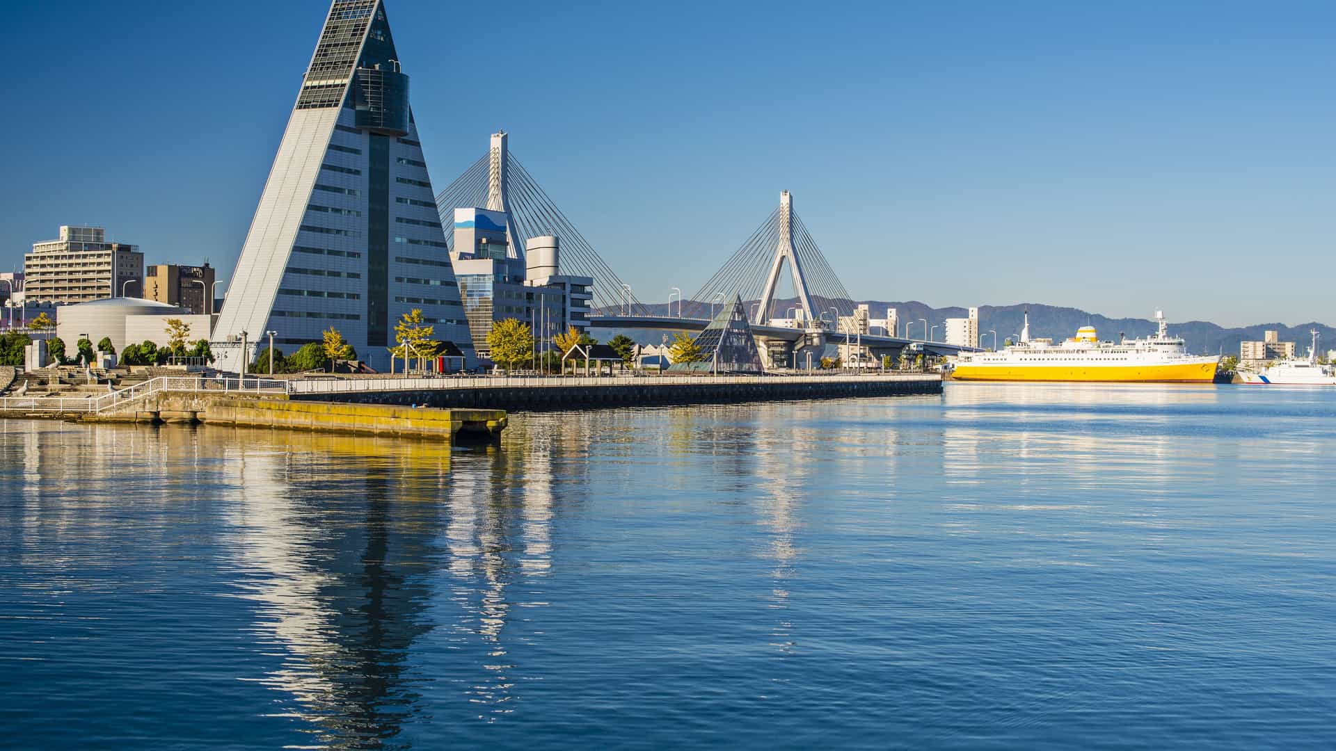 A wide, scenic view of the Aomori Bay Bridge and the iconic pyramid-shaped ASPAM building, with a large orange-and-white ferry docked in the harbor.