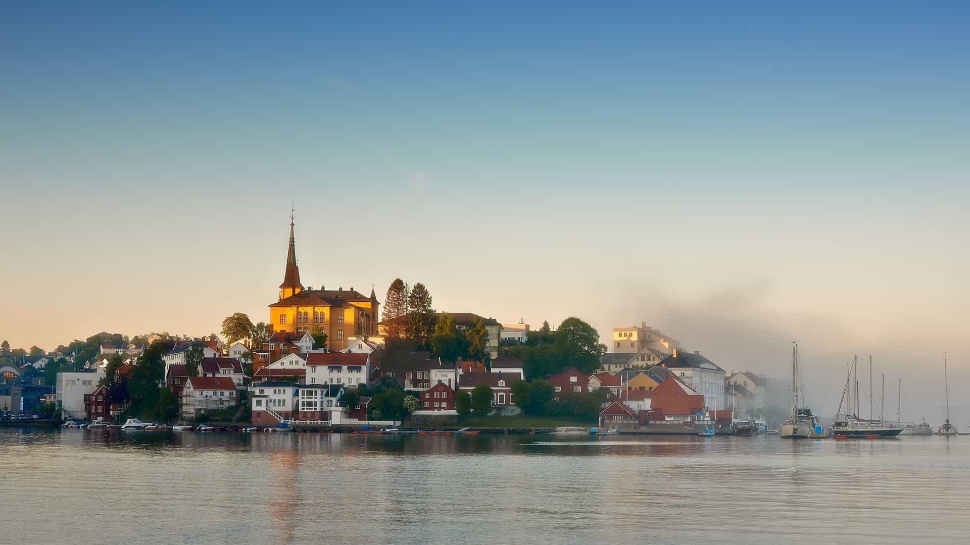 A stunning sunrise shot of the historic town of Arendal, Norway, with its church steeple and colorful buildings silhouetted against a golden sky and reflected in the calm fjord waters.