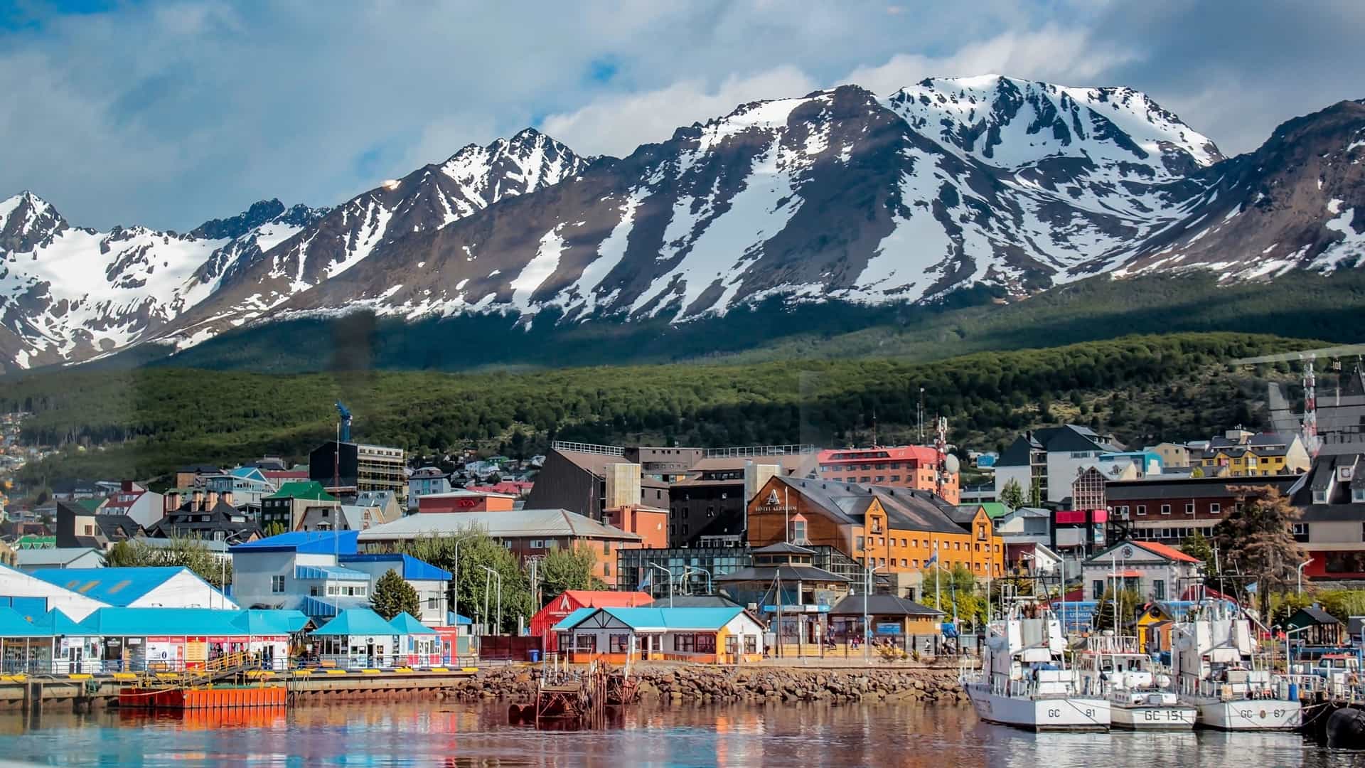 A wide-angle landscape of Ushuaia, Argentina, with a colorful waterfront town and a port in the foreground, set against a backdrop of the majestic, snow-capped Andes Mountains.