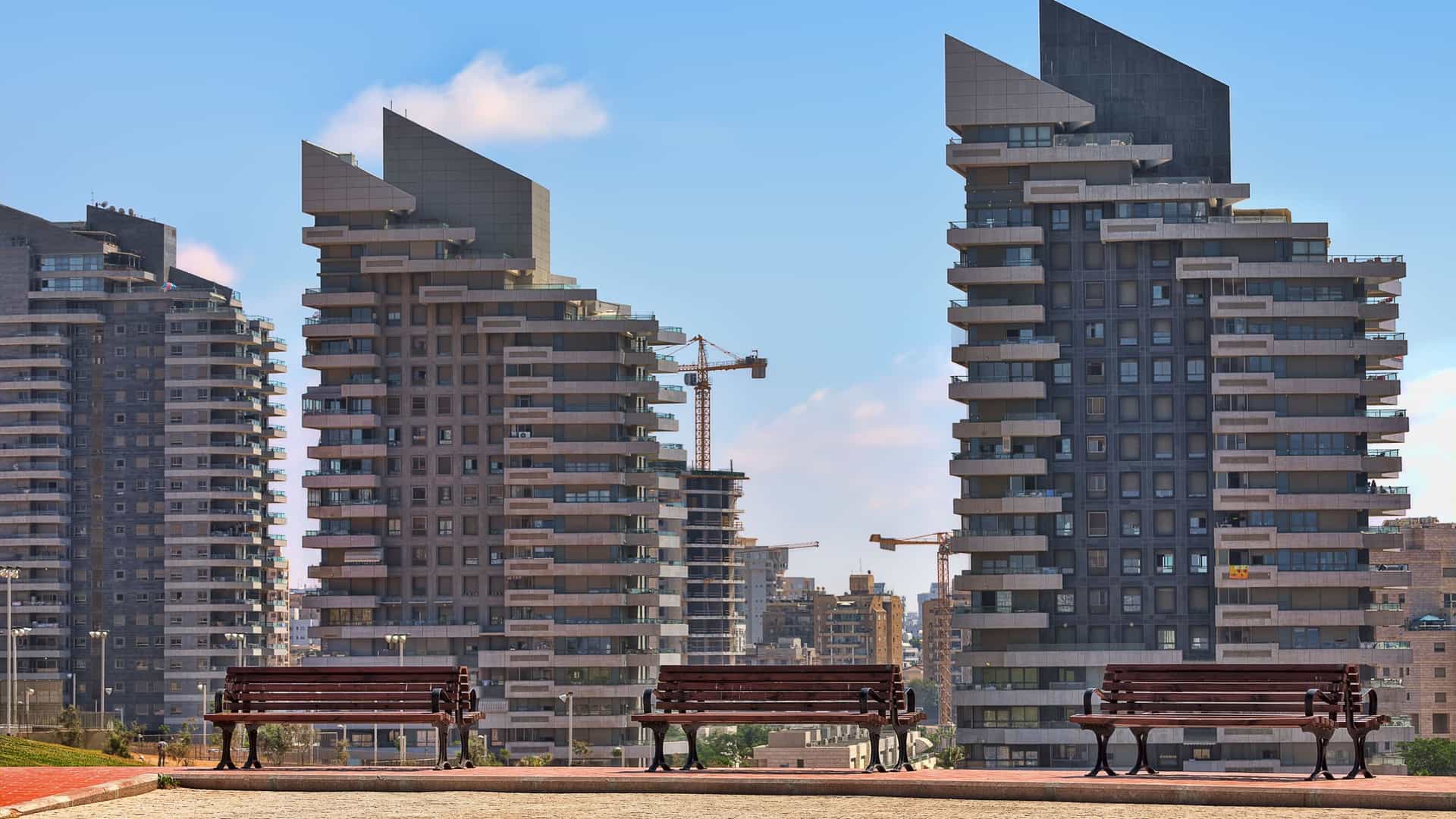 A scenic view of Ashdod, Israel, featuring modern, high-rise apartment buildings with unique architecture and three benches overlooking the urban landscape.