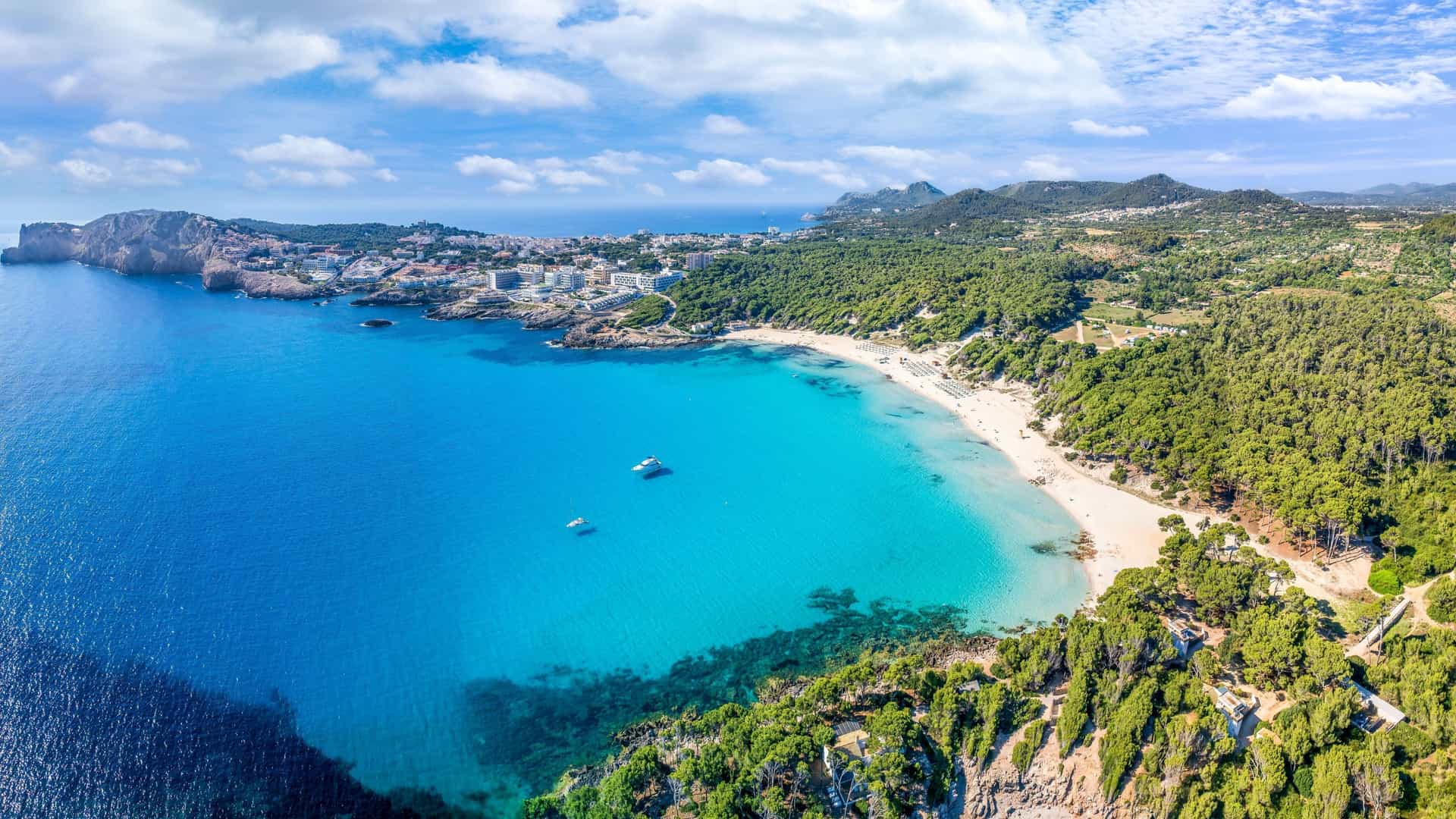 An aerial view of a picturesque beach on the Balearic Sea, with turquoise water and white sand framed by lush green trees and rocky cliffs.