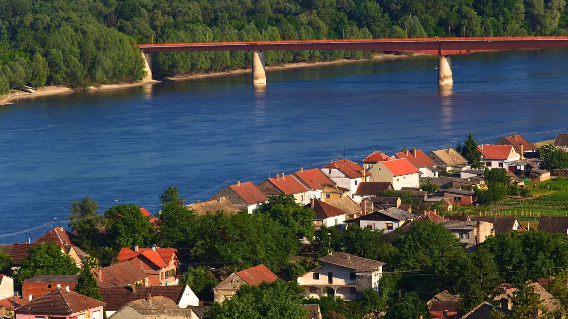An aerial view of a small town with red-roofed houses on the bank of a wide river, with a large red bridge connecting a forest in the background.