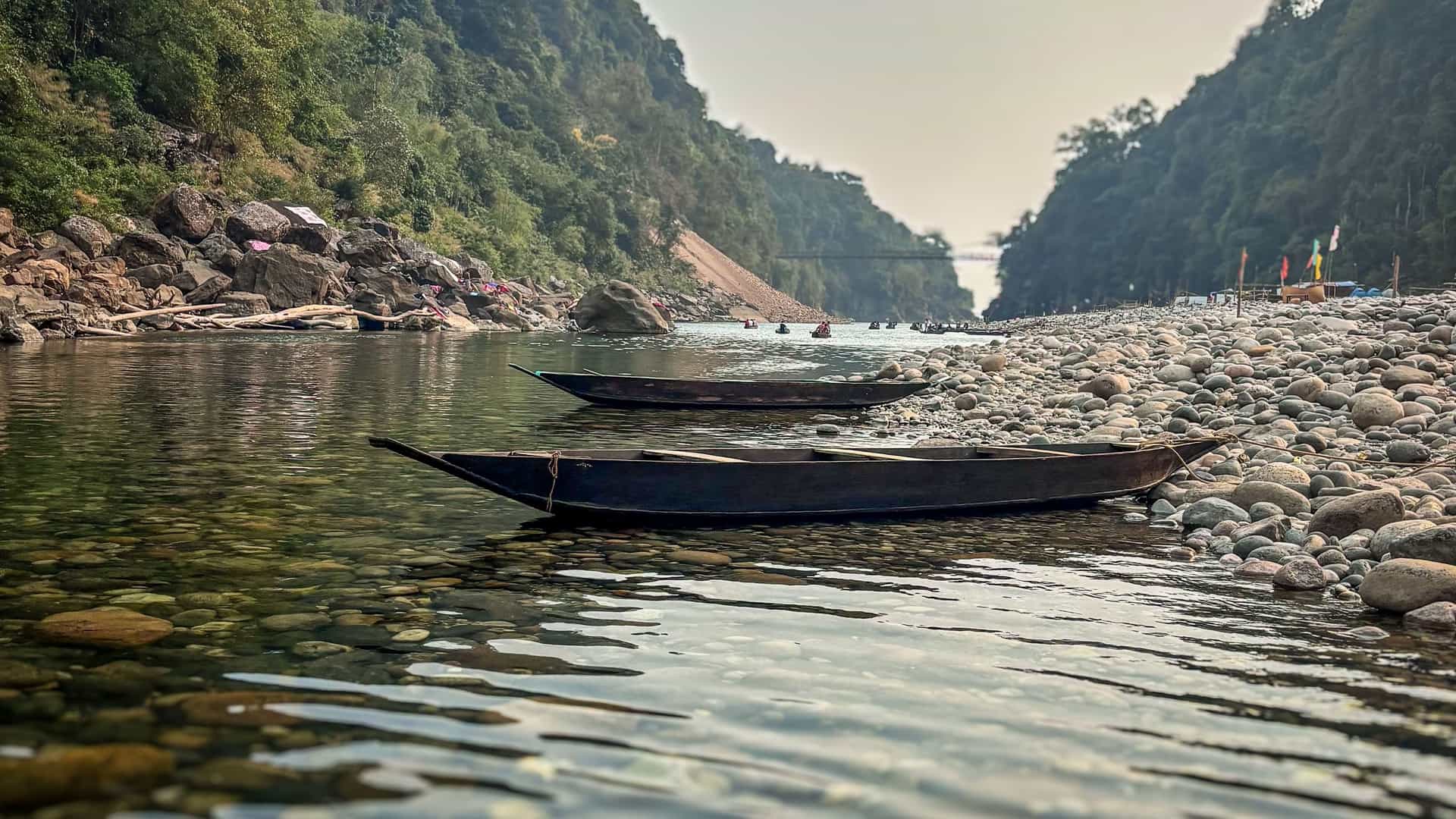 Two long, dark wooden boats resting on the clear, shallow water of a river that flows through a valley with rocky shores and lush green hillsides in the Bay of Bengal.